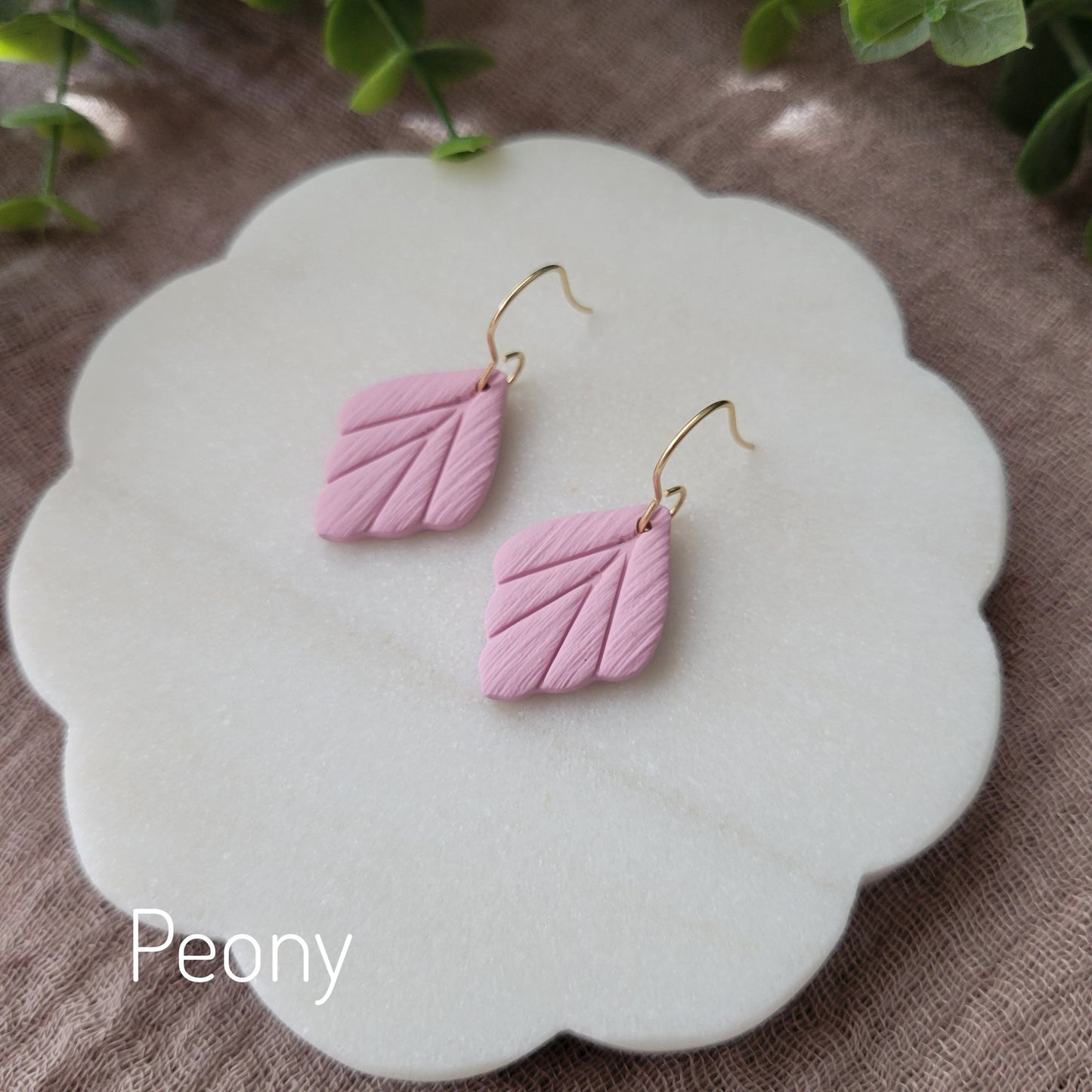 Side view of a pair of small pink leaf shaped earrings displayed on a white round scalloped tray.