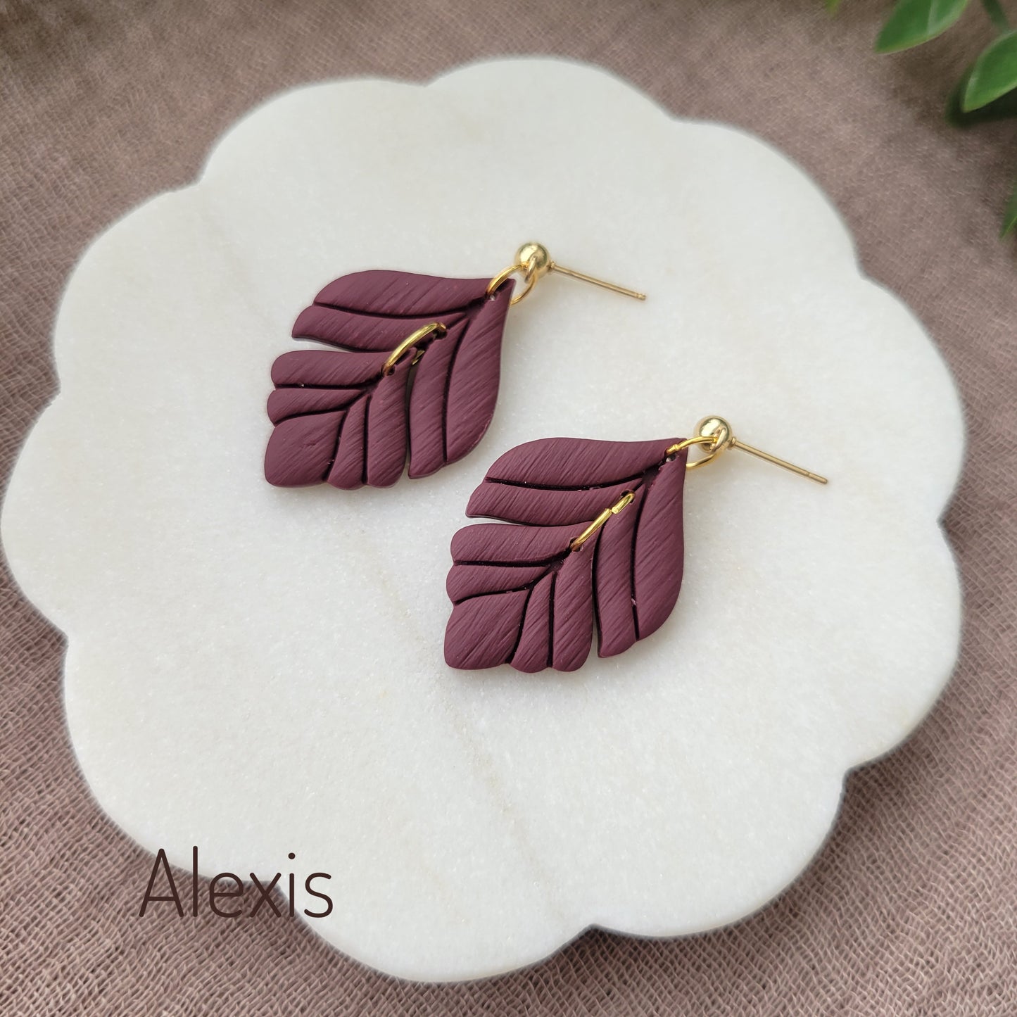Side view of a pair of reddish purple elegant leaf shaped earrings displayed on a white round scalloped tray.