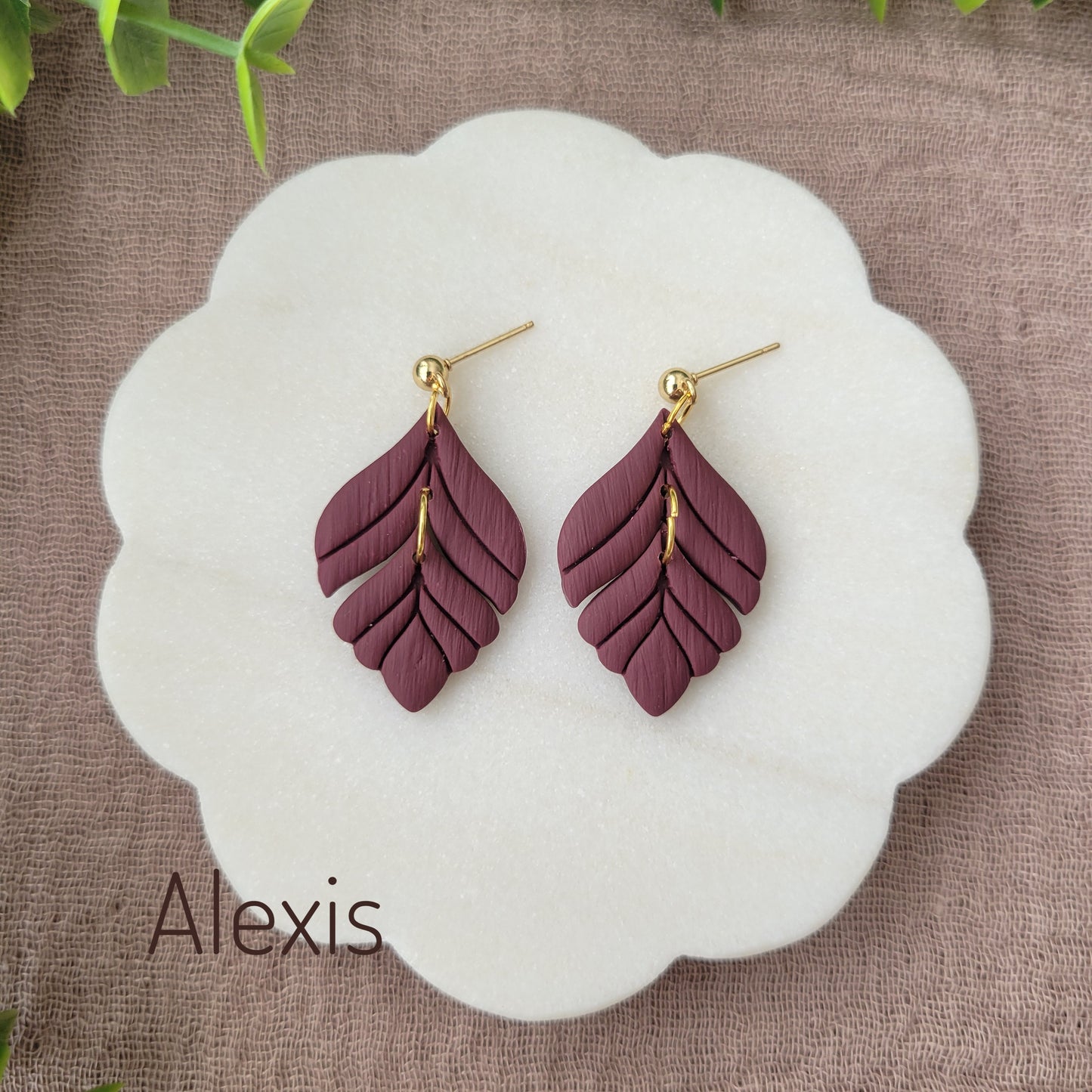 Top view of a pair of reddish purple elegant leaf shaped earrings displayed on a white round scalloped tray.