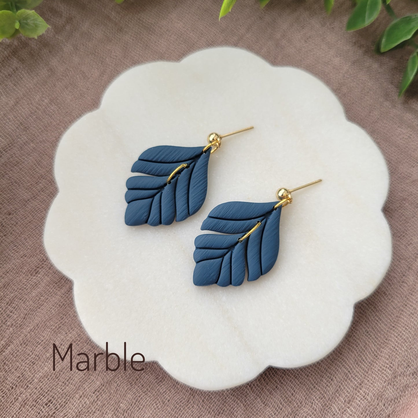 Side view of a pair of blue elegant leaf shaped earrings displayed on a white round scalloped tray.