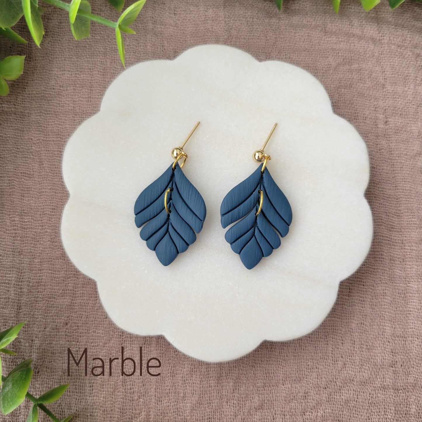 Top view of a pair of blue elegant leaf shaped earrings displayed on a white round scalloped tray.
