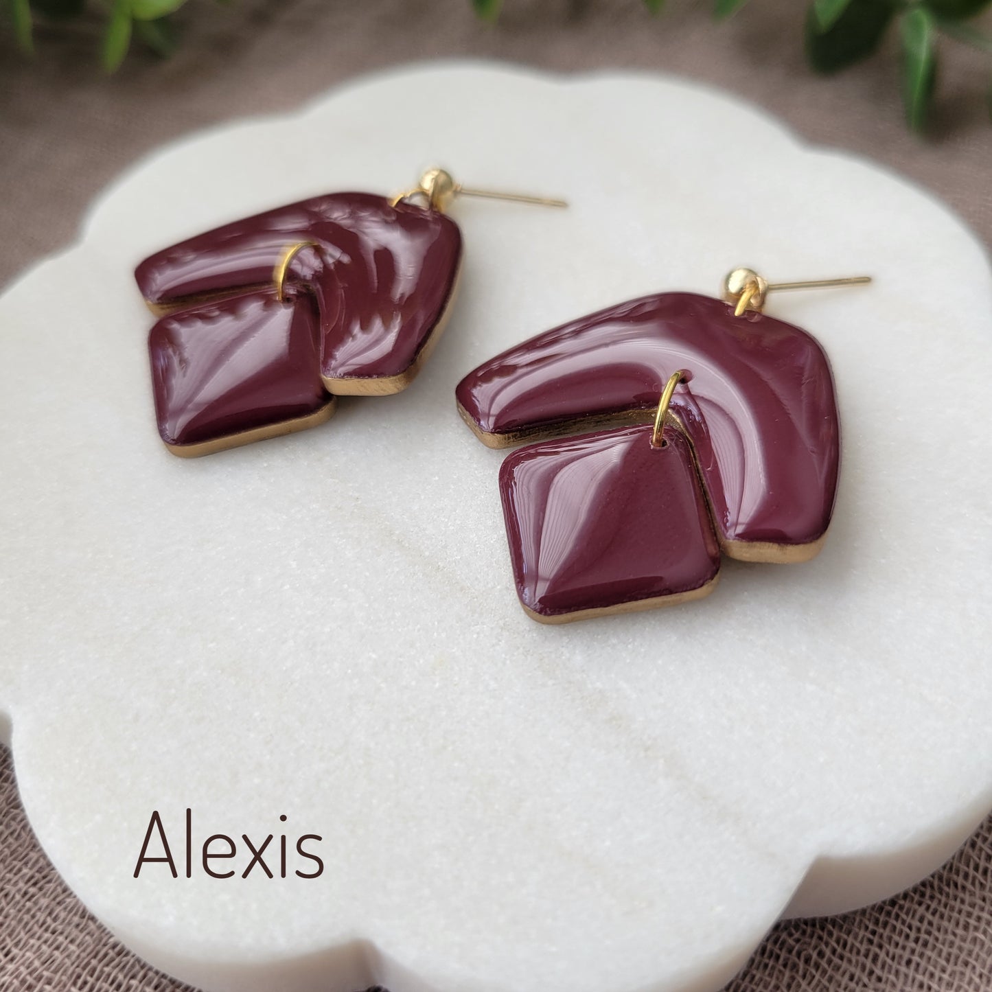 Side view of a pair of reddish purple earrings with shinny resin displayed on a white round scalloped tray.