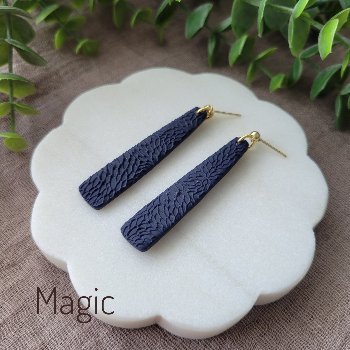 Side view of a pair of dark blue pillar earrings with interwoven leaves texture displayed on a white round scalloped tray.