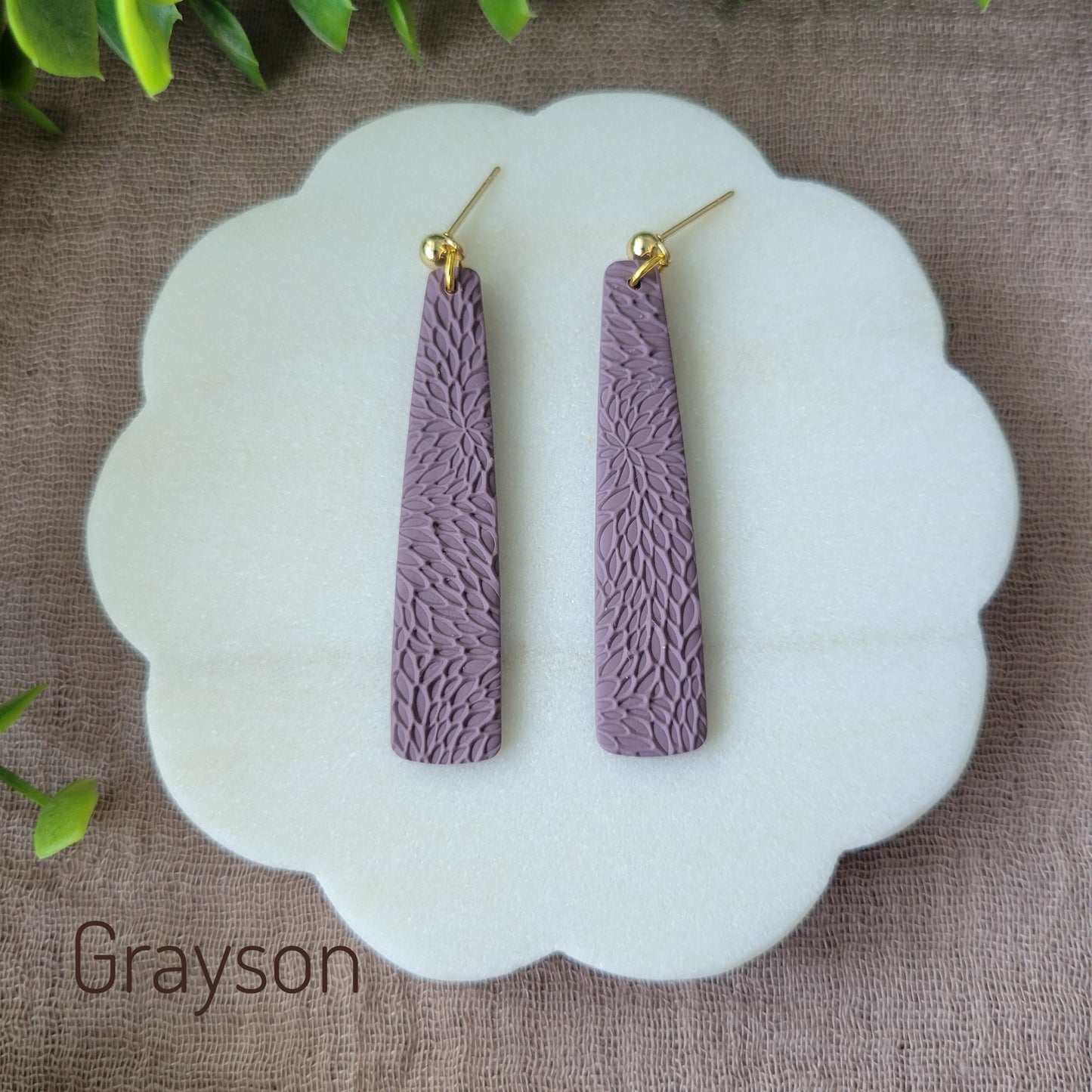Top view of a pair of light purple pillar earrings with interwoven leaves texture displayed on a round white scalloped tray.