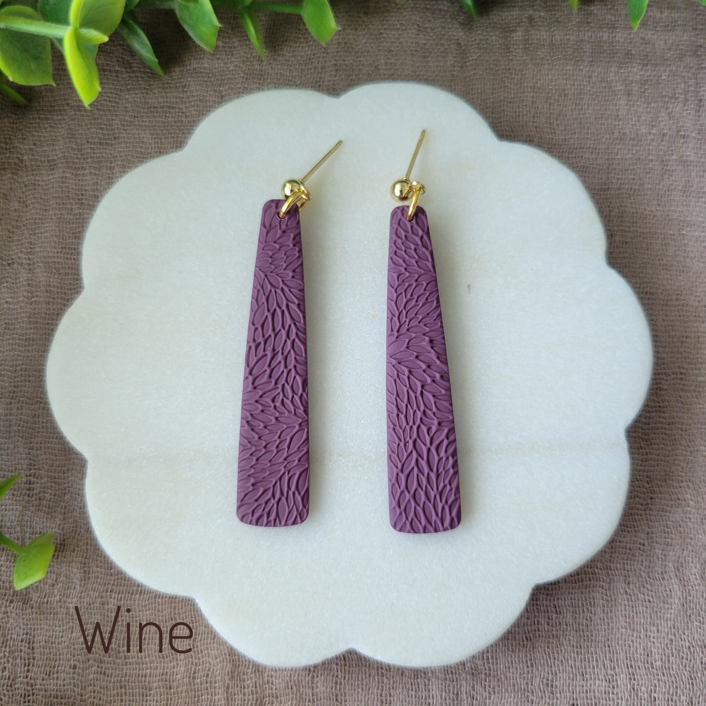 Top view of a pair of reddish purple pillar earrings with interwoven leaves texture displayed on a white round scalloped tray.
