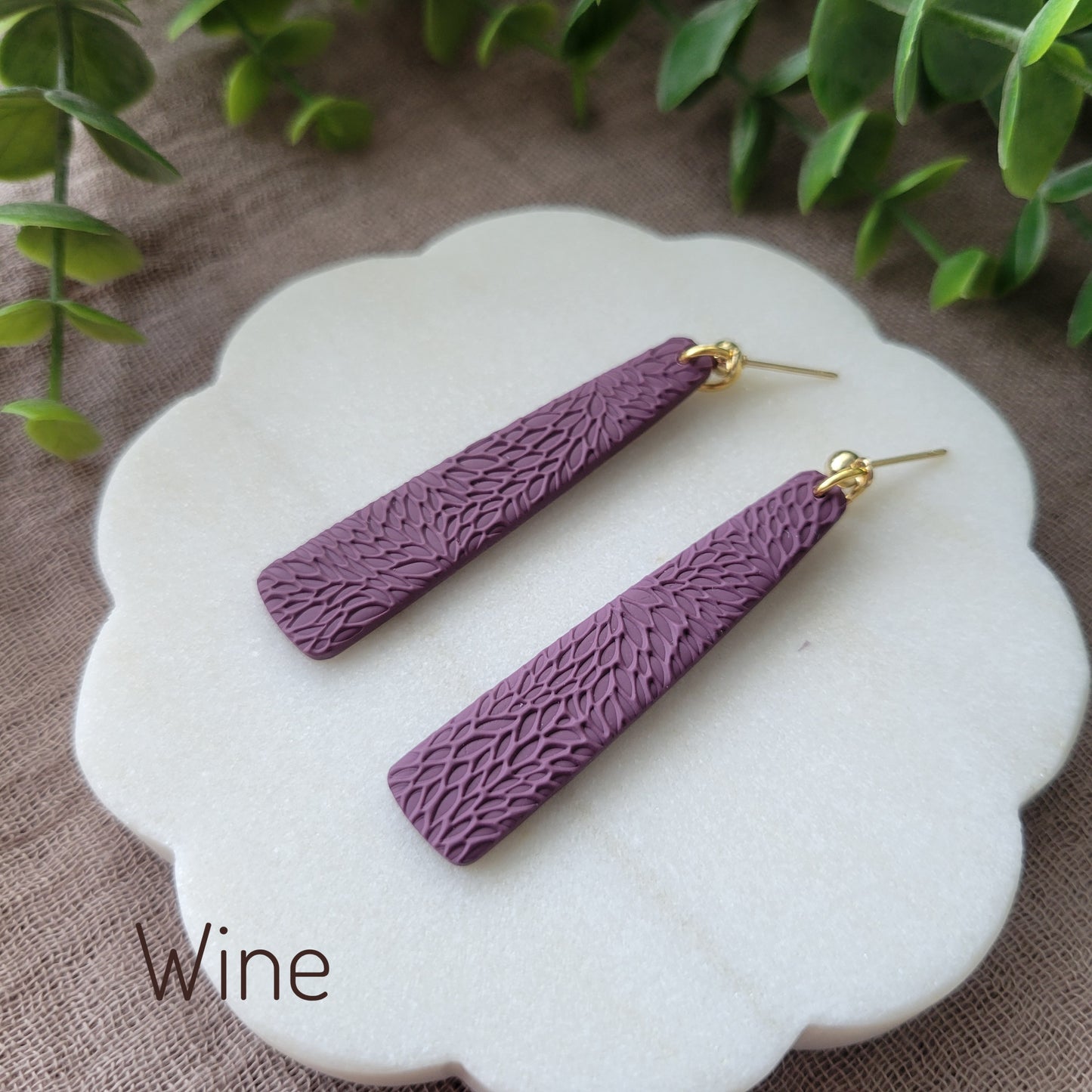 Side view of a pair of reddish purple pillar earrings with interwoven leaves texture displayed on a white round scalloped tray.