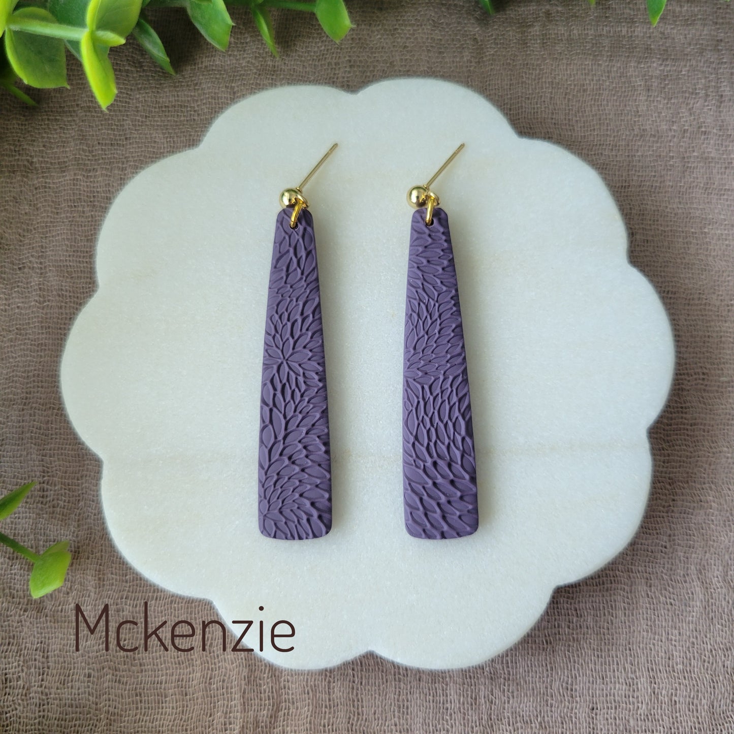 Top view of a pair of dark purple pillar earrings with interwoven leaves texture displayed on a white round scalloped tray.