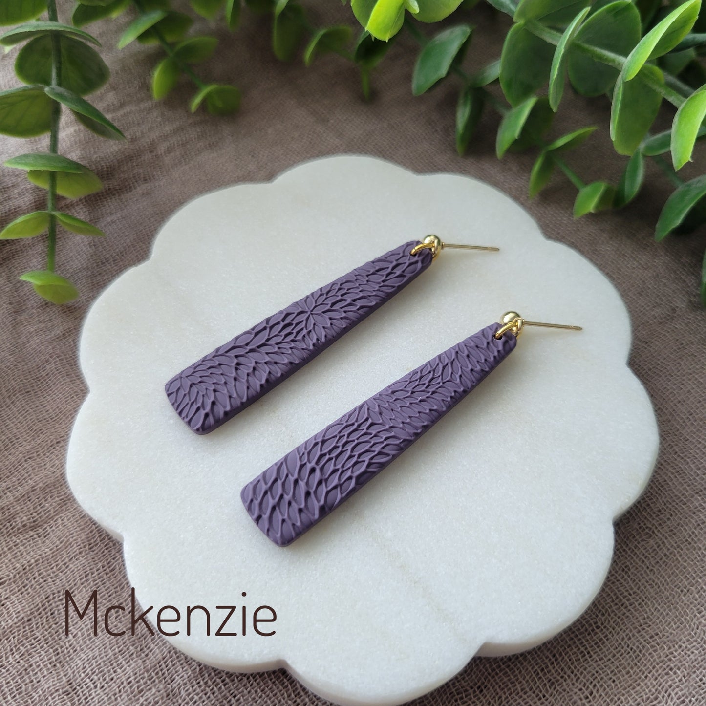 Side view of a pair of dark purple pillar earrings with interwoven leaves texture displayed on a white round scalloped tray.
