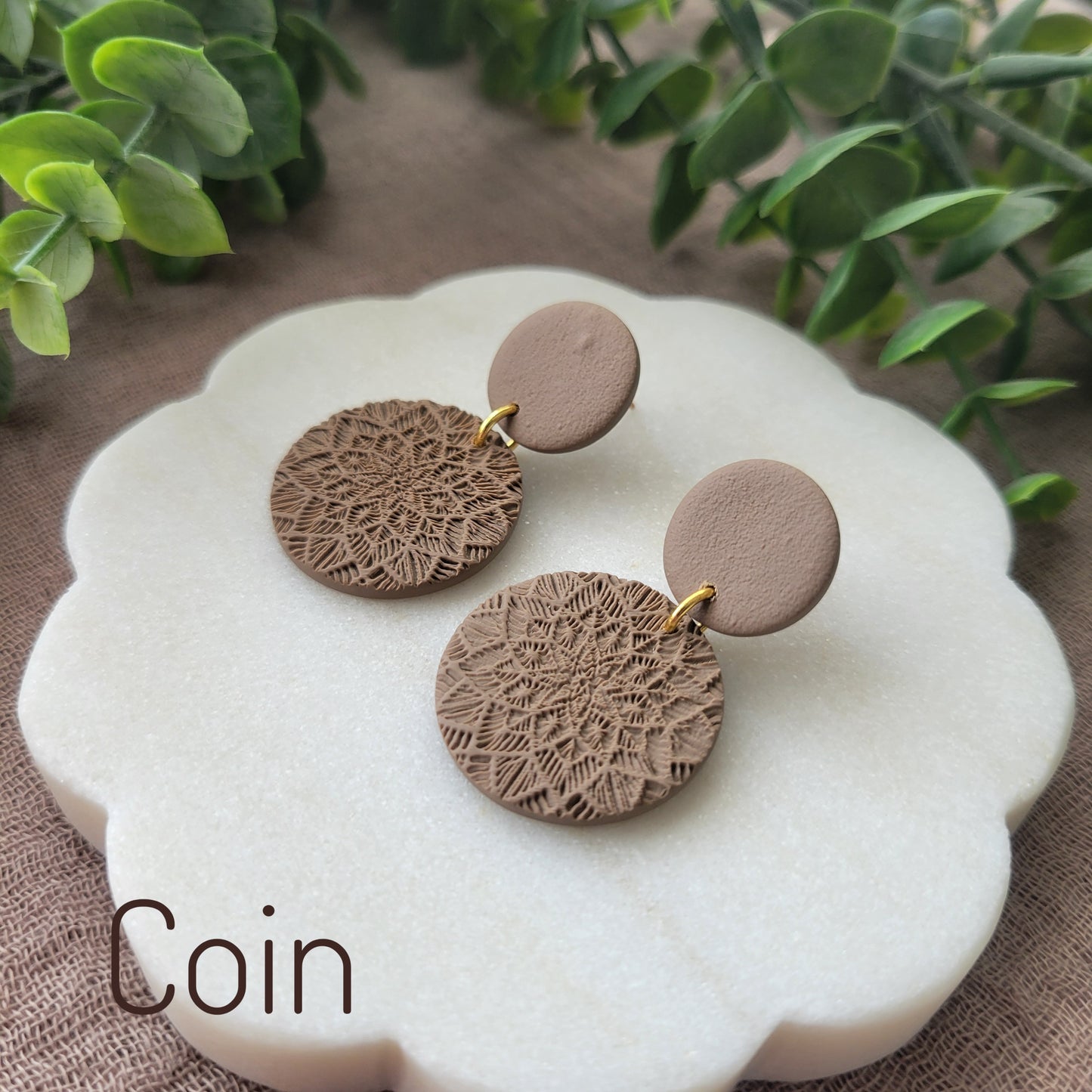 Side view of a pair of brown mandala textured earrings displayed on a white round scalloped tray.