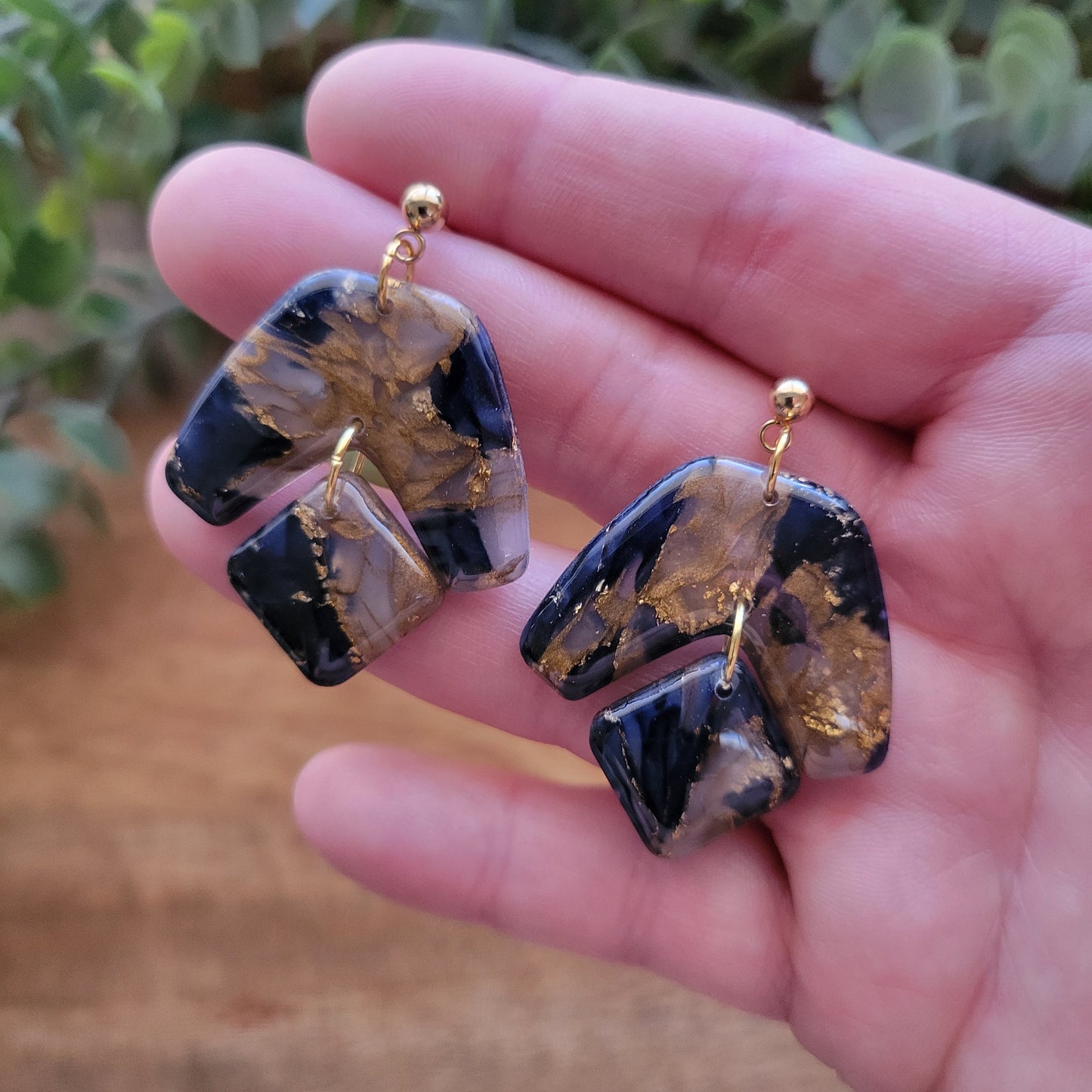 Close up top view of a pair of dark blue, bronze marble boomerang shaped earrings displayed on a hand.
