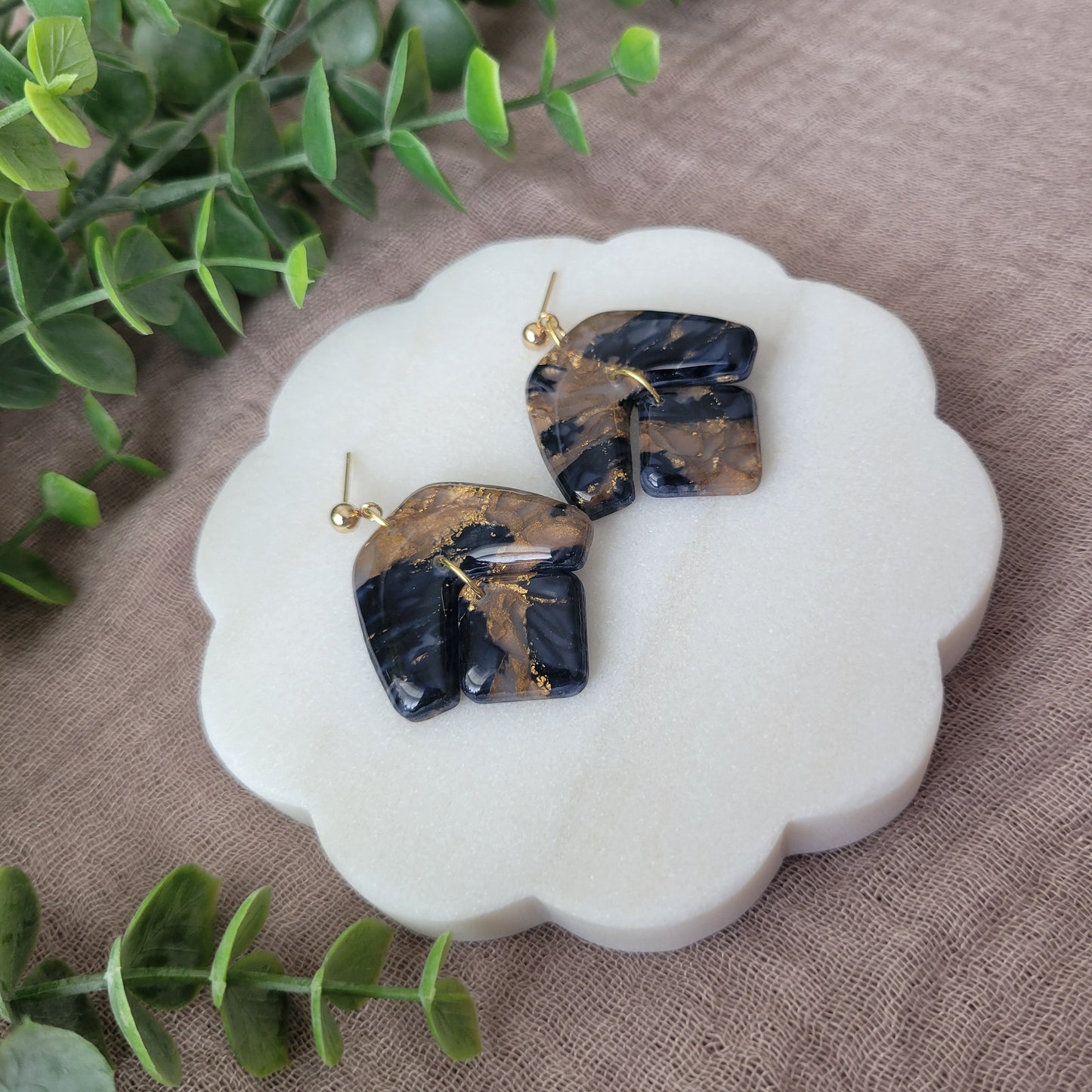 Left side view of a pair of dark blue, bronze marble boomerang shaped earrings displayed on a white round scalloped tray.