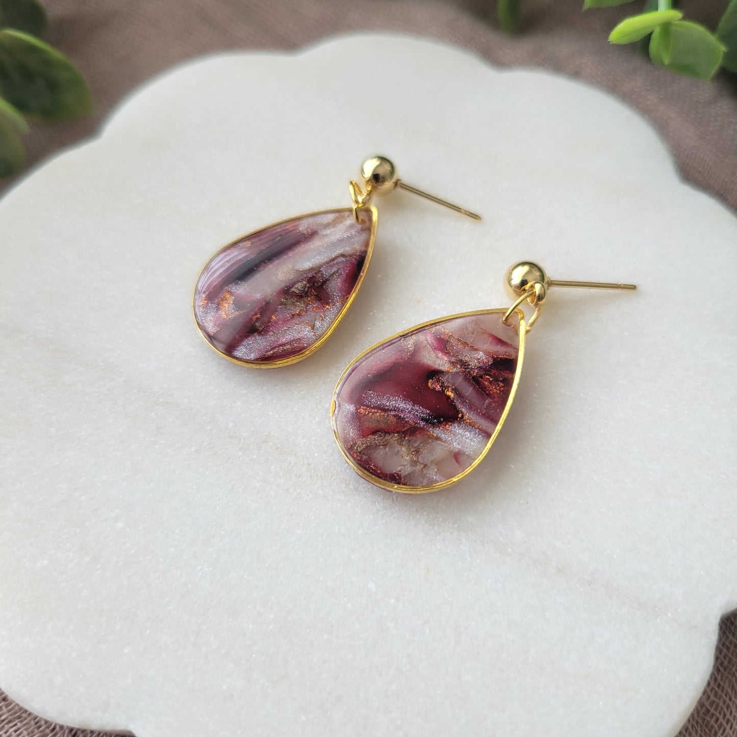Up close right side view of a pair of maroon, pearl teardrop earrings with a gold trim bezel displayed on a white round scalloped tray.