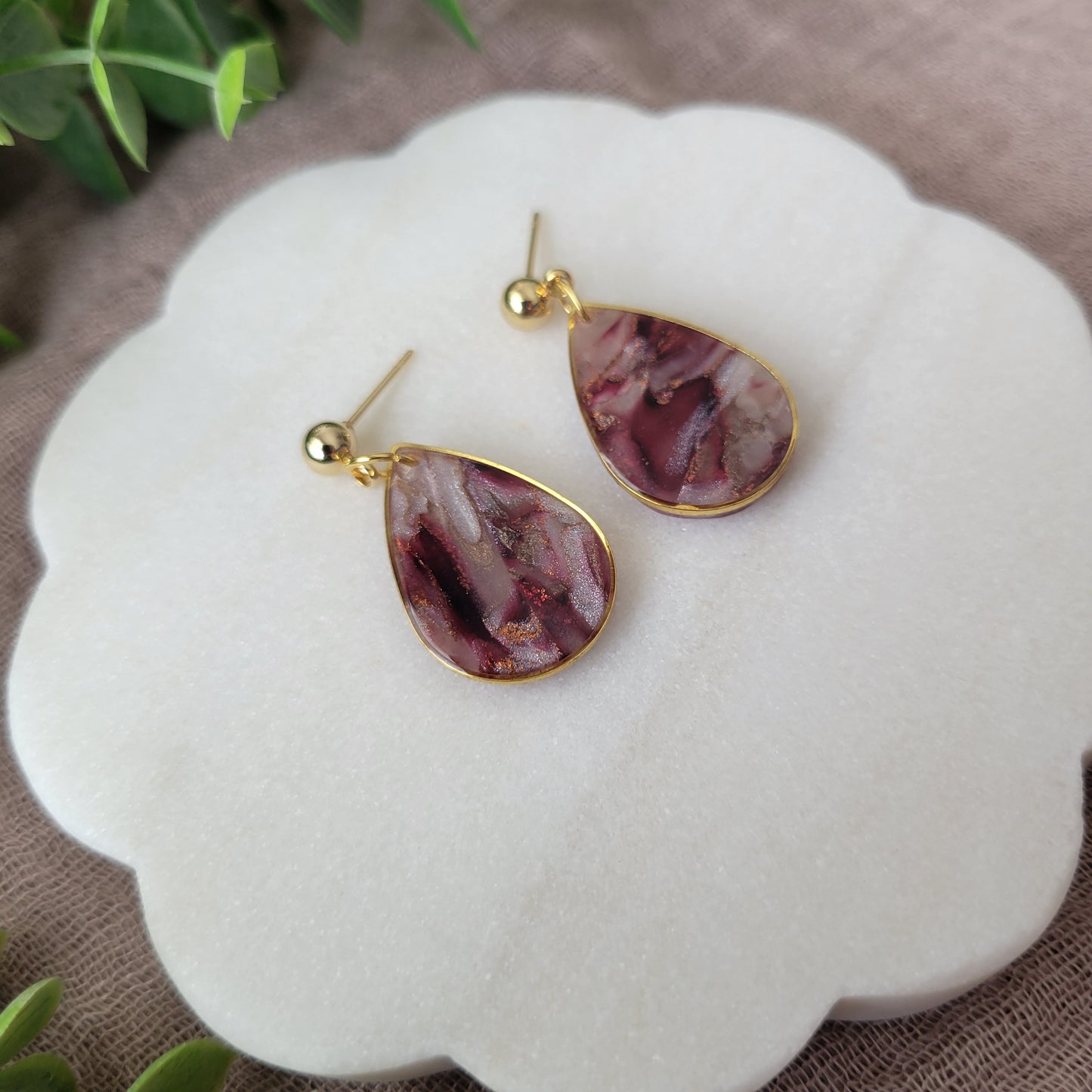 Left side view of a pair of maroon, pearl teardrop earrings with a gold trim bezel displayed on a white round scalloped tray.