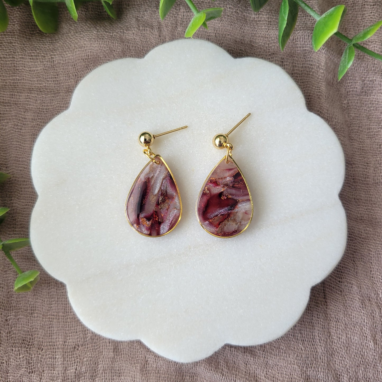 Top view of a pair of maroon, pearl teardrop earrings with a gold trim bezel displayed on a white round scalloped tray.