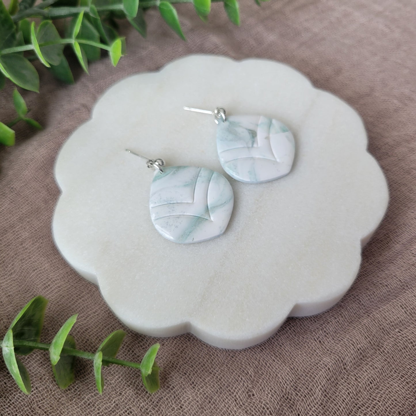Left side view of a pair of tiffany blue, white marble embossed round tear drop shaped earrings displayed on a white round scalloped tray.