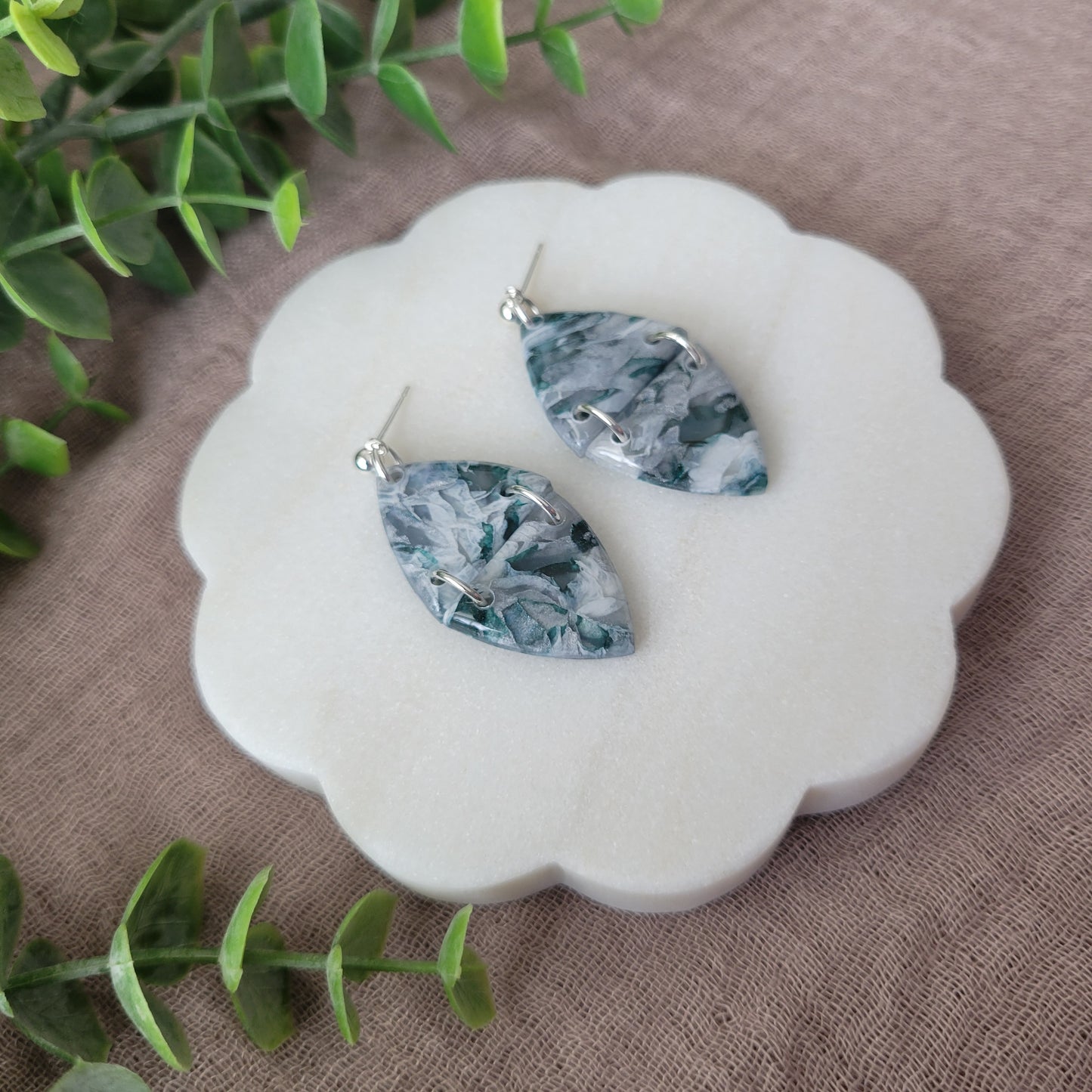 Left side view of a pair of green, white, silver marble earrings displayed on a white round scalloped tray.