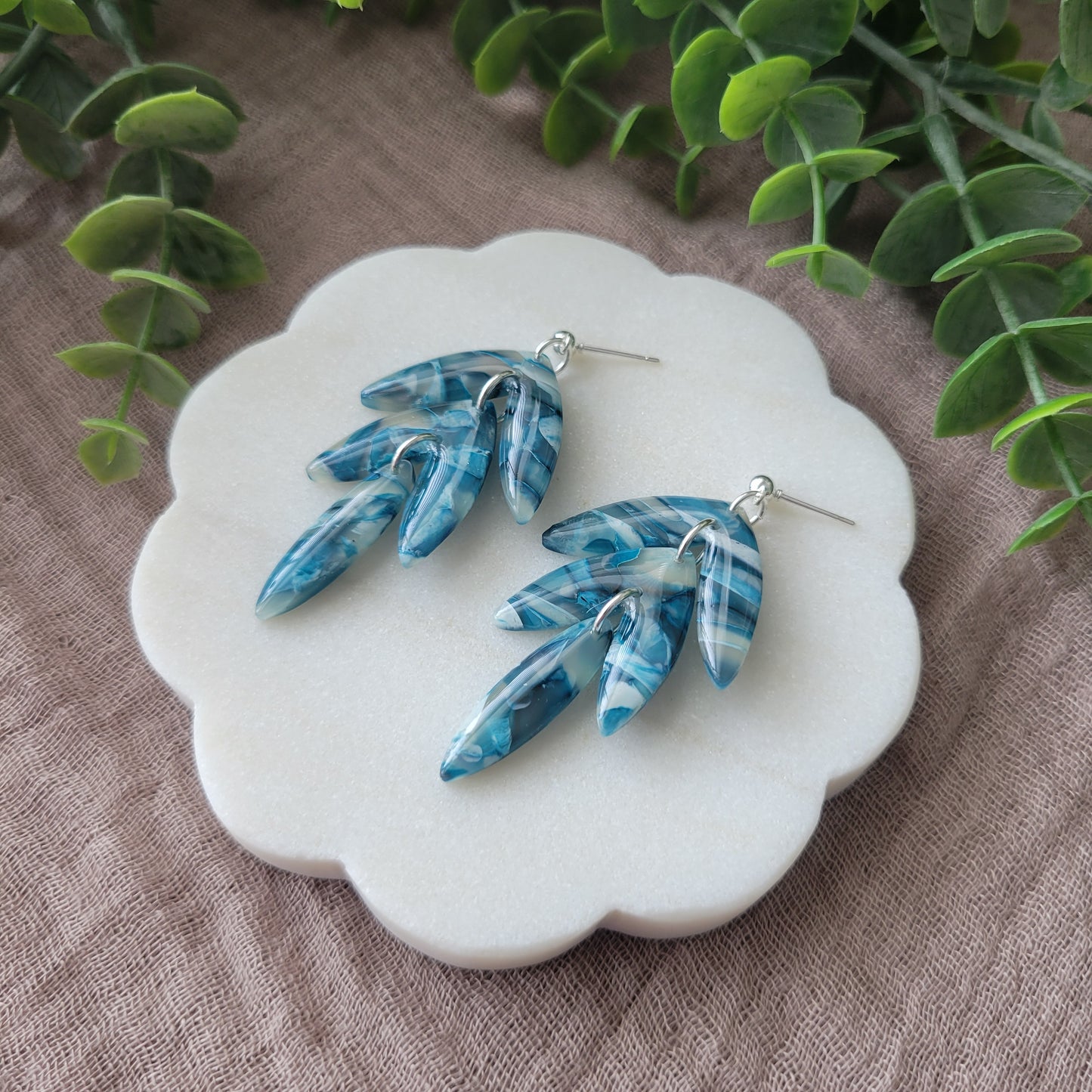 Right side view of a pair of blue marble long leaf shaped earrings displayed on a white round scalloped tray.
