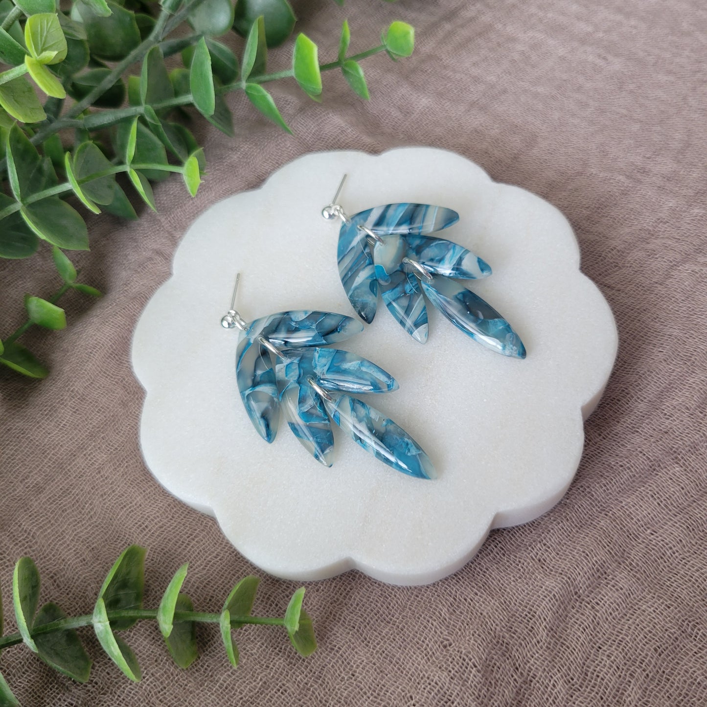 Left side view of a pair of blue marble long leaf shaped earrings displayed on a white round scalloped tray.
