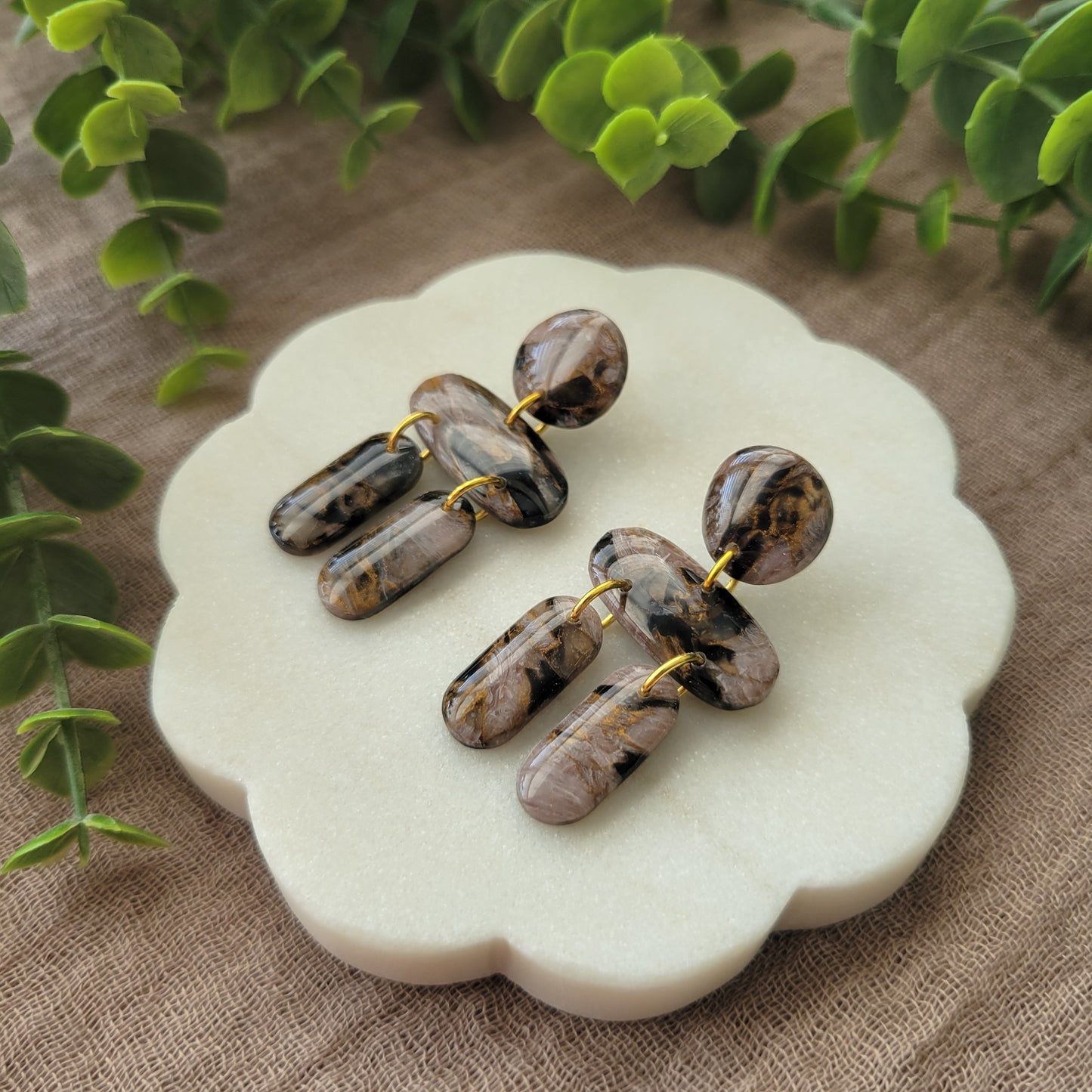 Right side view of a pair of brown, black, gold accented stone shaped earrings displayed on a white round scalloped tray.