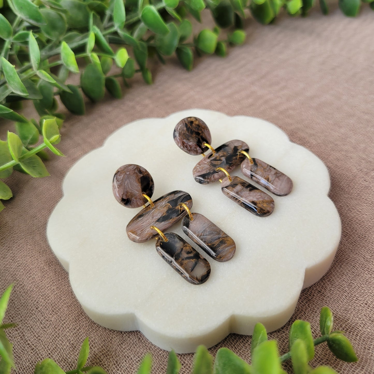 Left side view of a pair of brown, black, gold accented stone shaped earrings displayed on a white round scalloped tray.