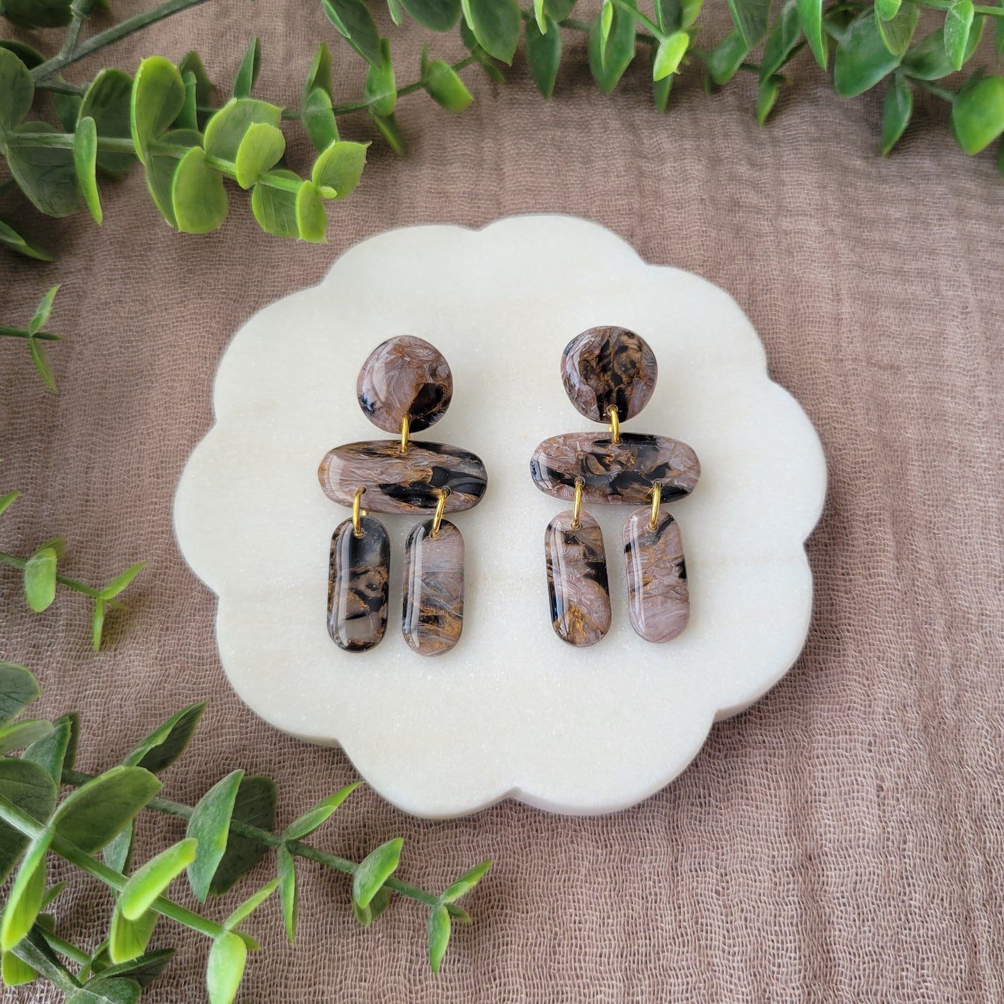 Top view of a pair of brown, black, gold accented stone shaped earrings displayed on a white round scalloped tray.