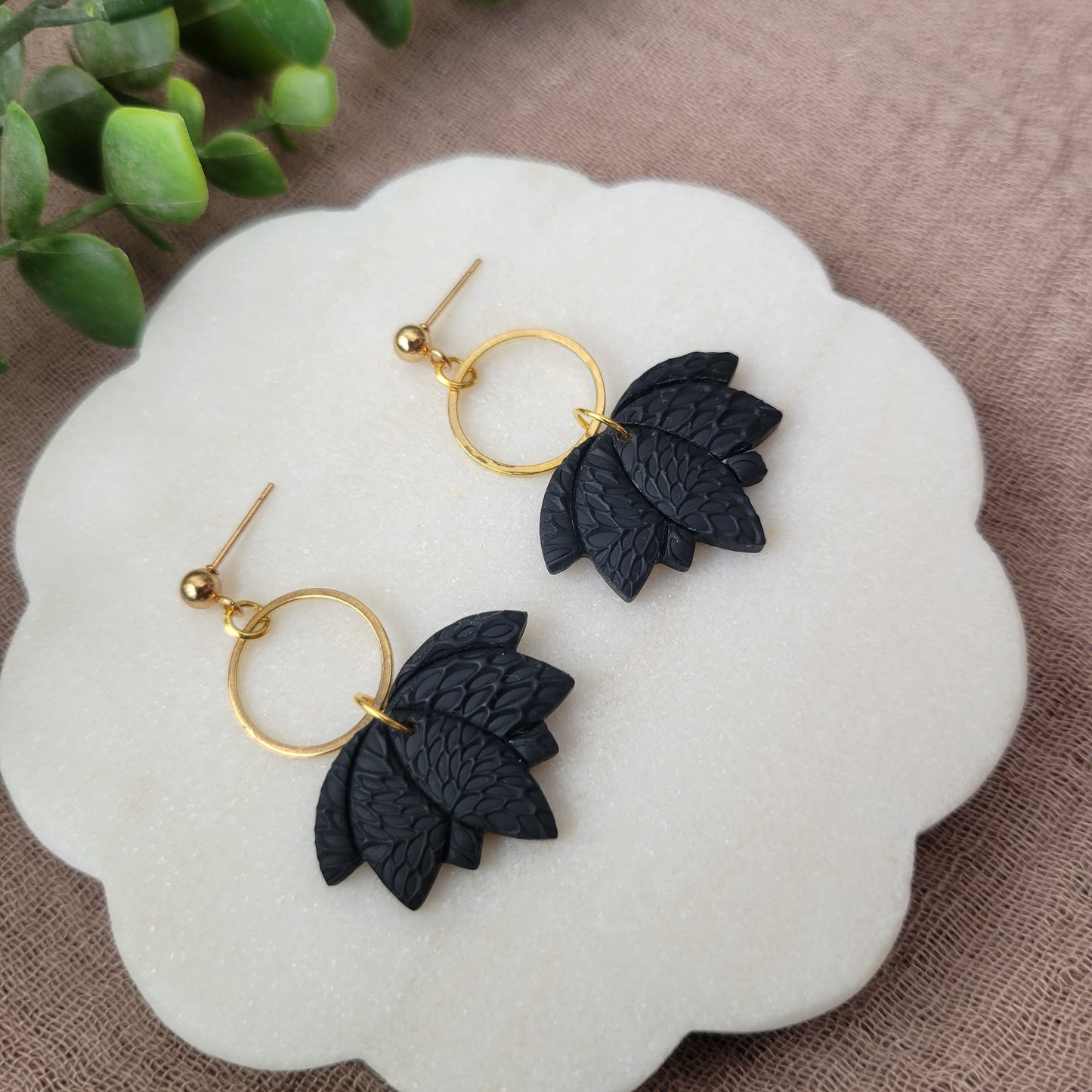 Left side view of a pair of black lotus earrings with interwoven leaves texture and gold circle bezel displayed on a white round scalloped tray.