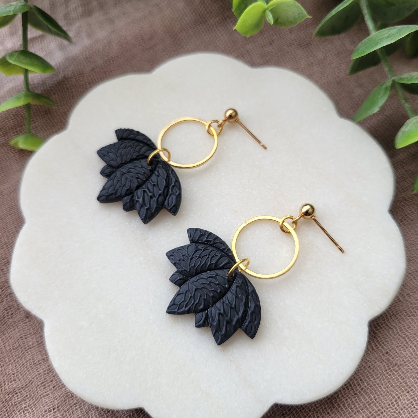 Right side view of a pair of black lotus earrings with interwoven leaves texture and gold circle bezel displayed on a white round scalloped tray.