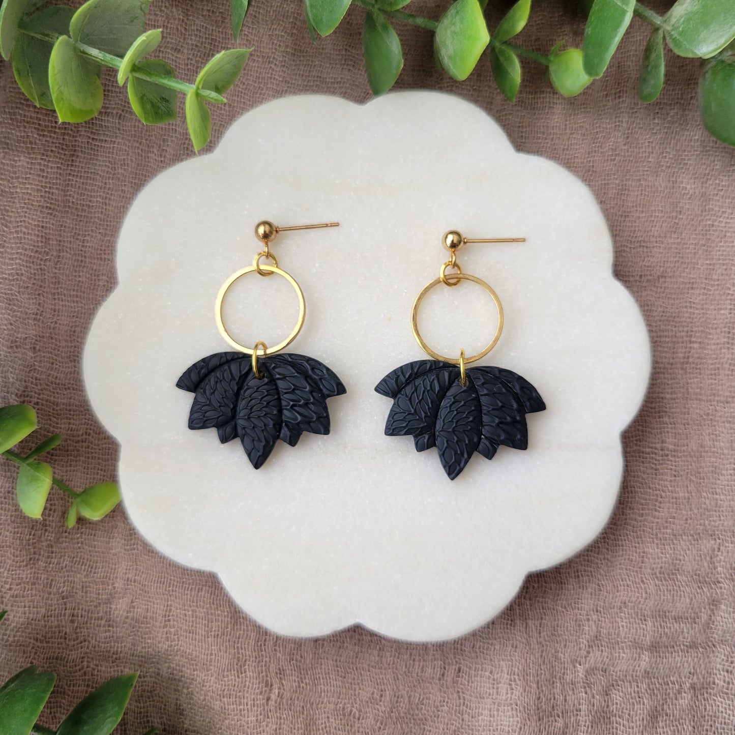 Top view of a pair of black lotus earrings with interwoven leaves texture and gold circle bezel displayed on a white round scalloped tray.