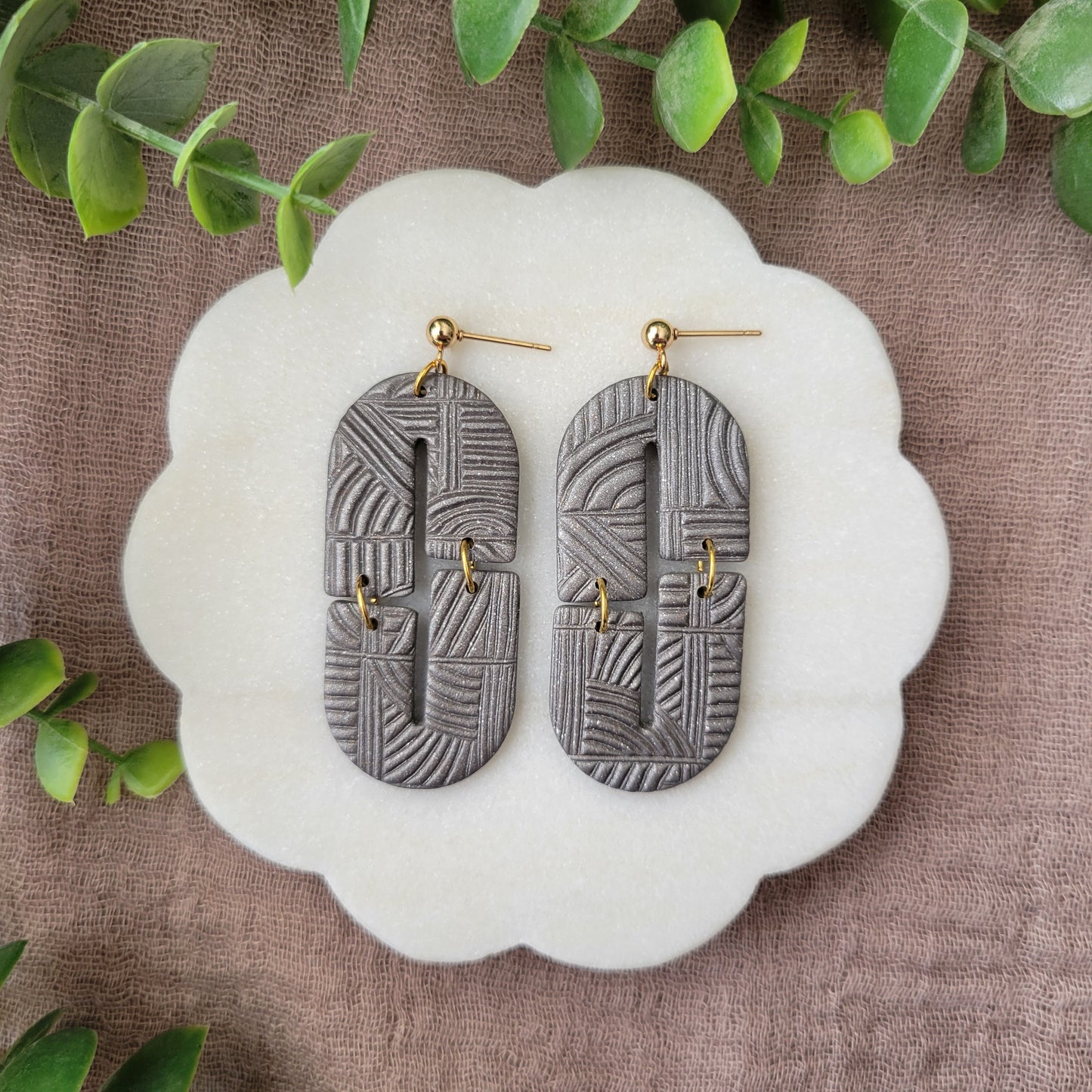 Top view of a pair of gray earrings with art deco lines texture displayed on a white round scalloped tray.