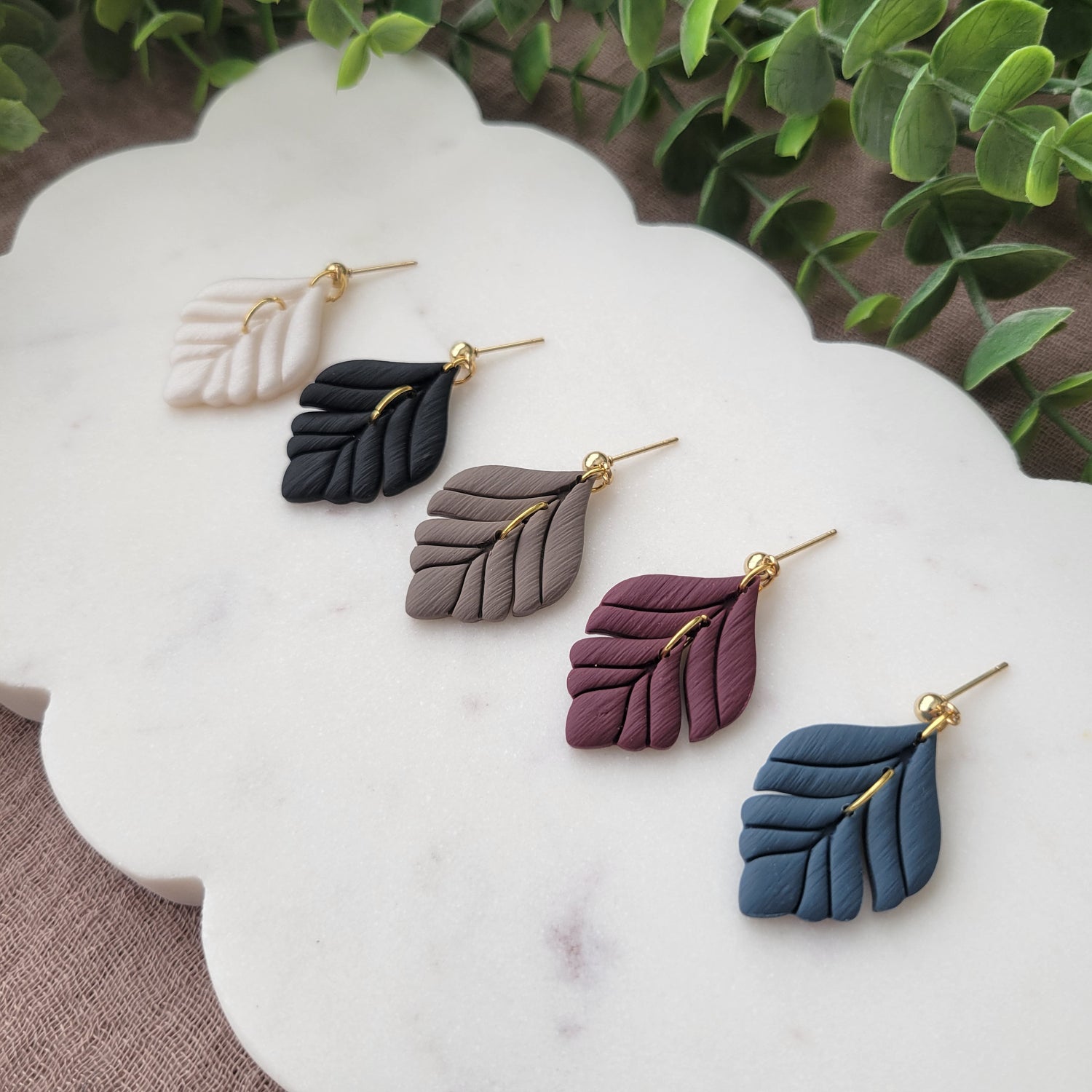 Side view of a set of five polymer clay elegant leaf shaped earrings displayed on a white scalloped tray.