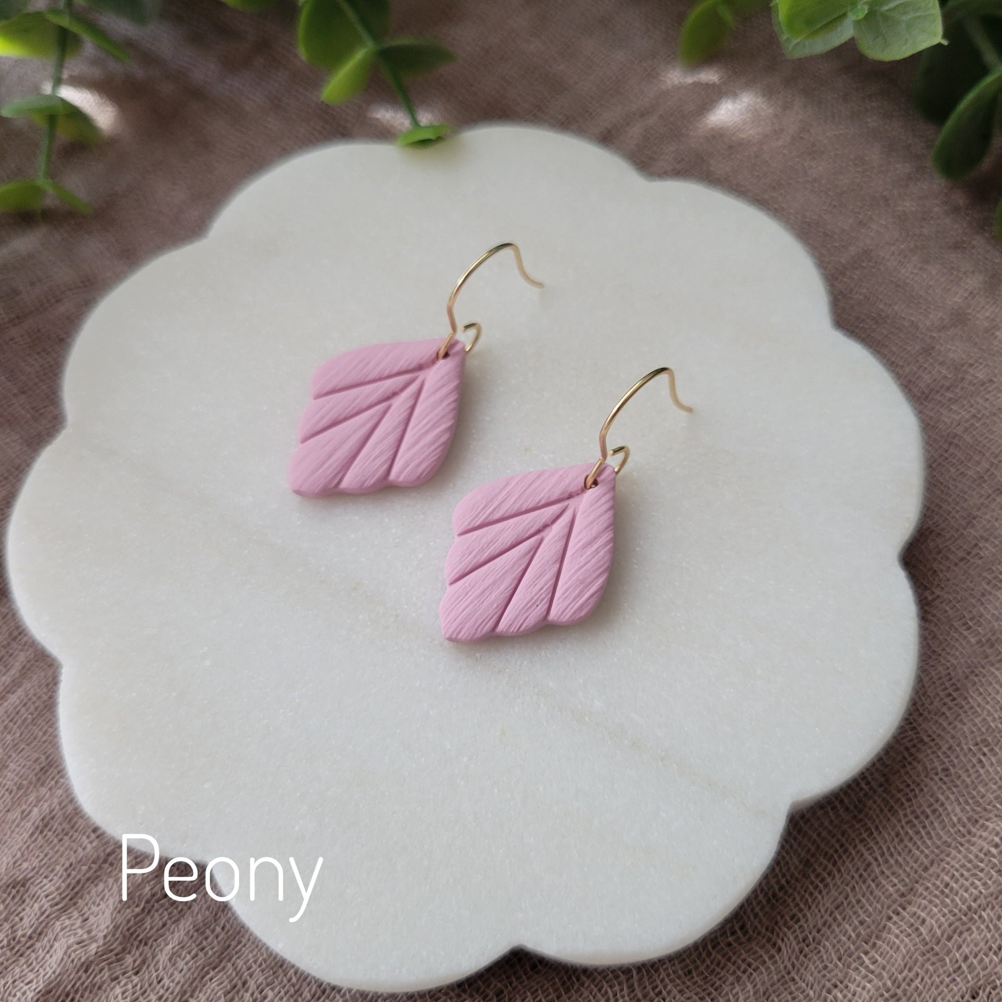 Side view of a pair of small pink leaf shaped earrings displayed on a white round scalloped tray.