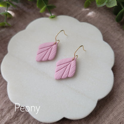 Side view of a pair of small pink leaf shaped earrings displayed on a white round scalloped tray.