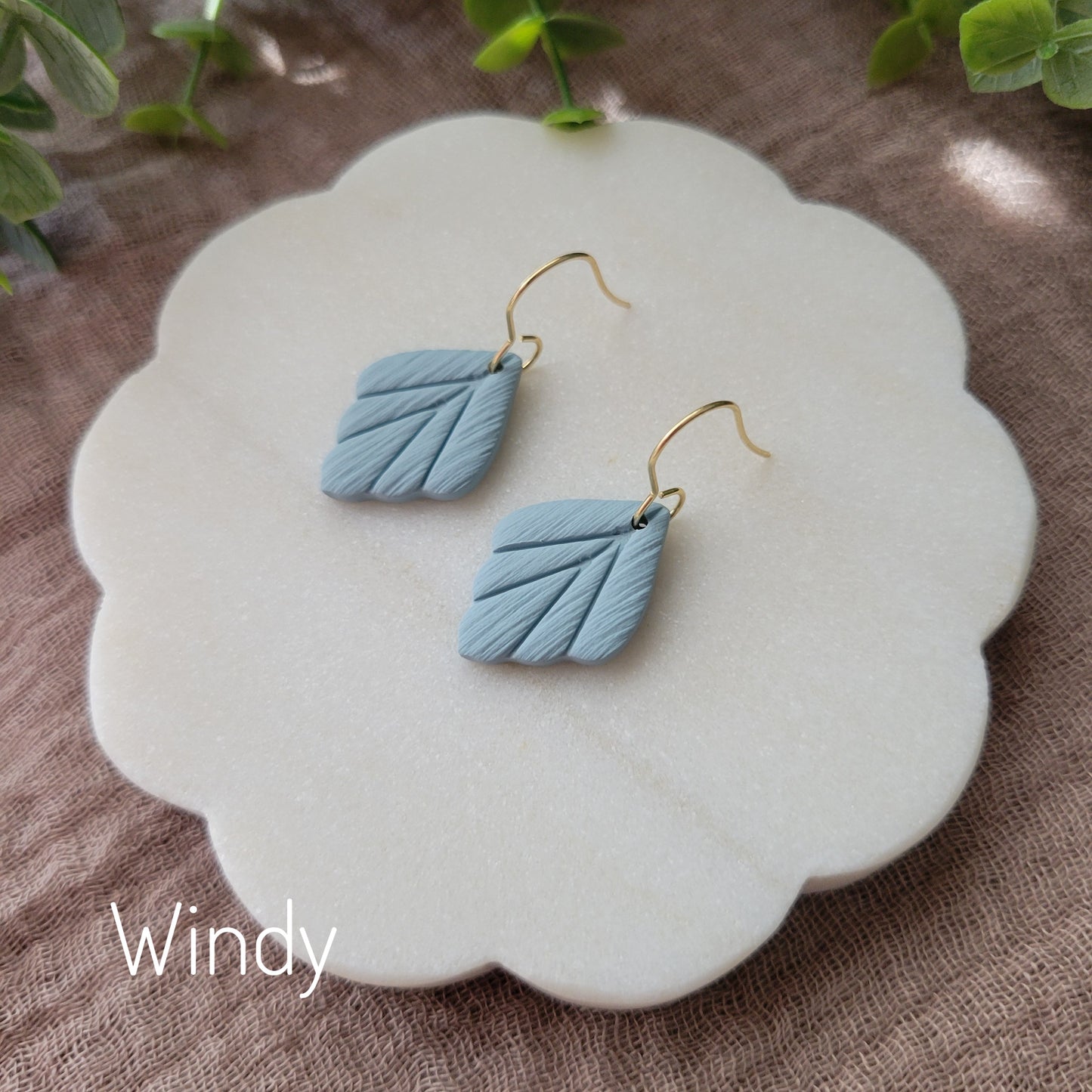 Side view of a pair of small blue leaf shaped earrings displayed on a white round scalloped tray.