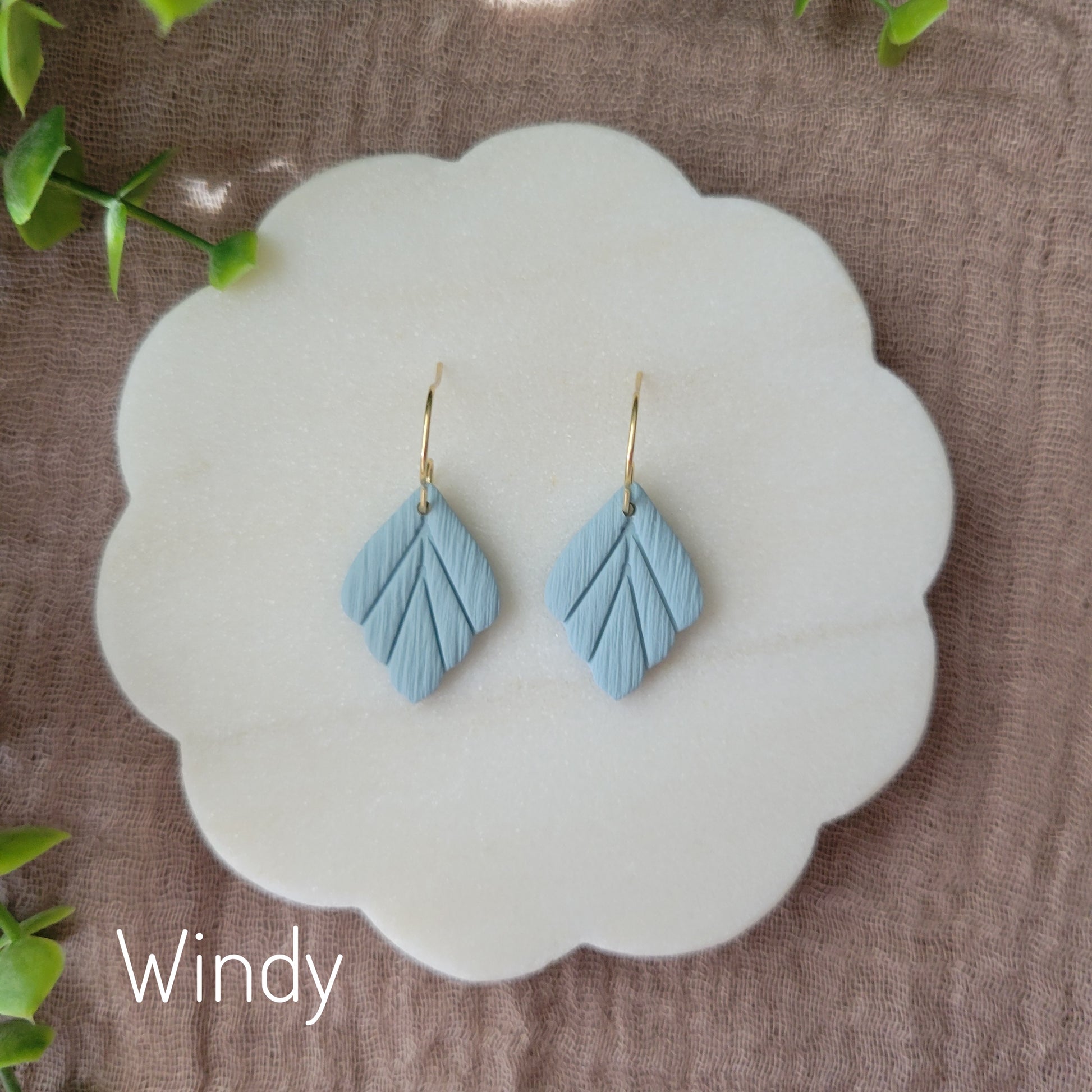 Top view of a pair of small blue leaf shaped earrings displayed on a white round scalloped tray.