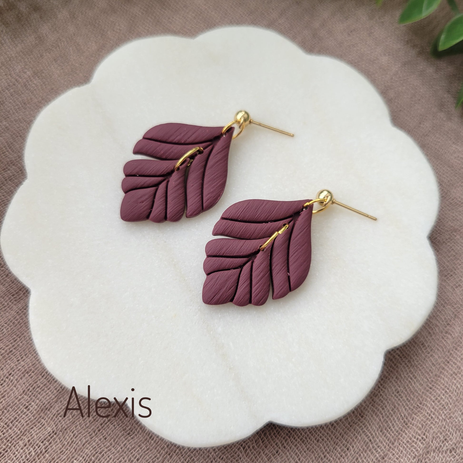 Side view of a pair of reddish purple elegant leaf shaped earrings displayed on a white round scalloped tray.