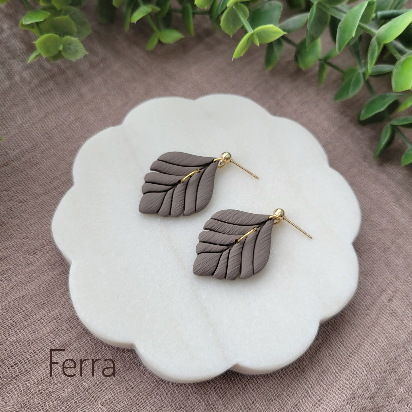 Side view of a pair of grayish brown elegant leaf shaped earrings displayed on a white round scalloped tray.