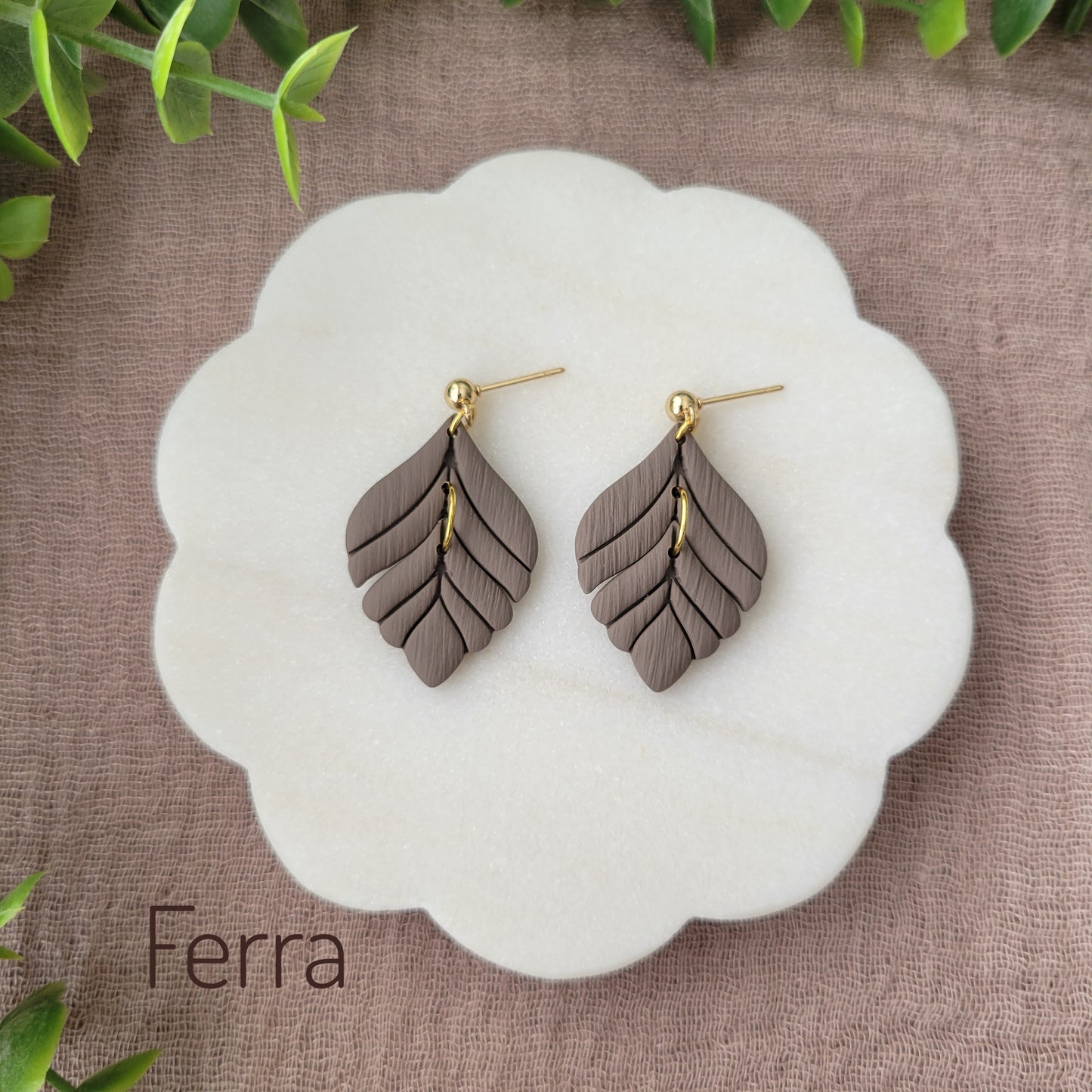 Top view of a pair of greyish brown elegant leaf shaped earrings displayed on a white round scalloped tray.