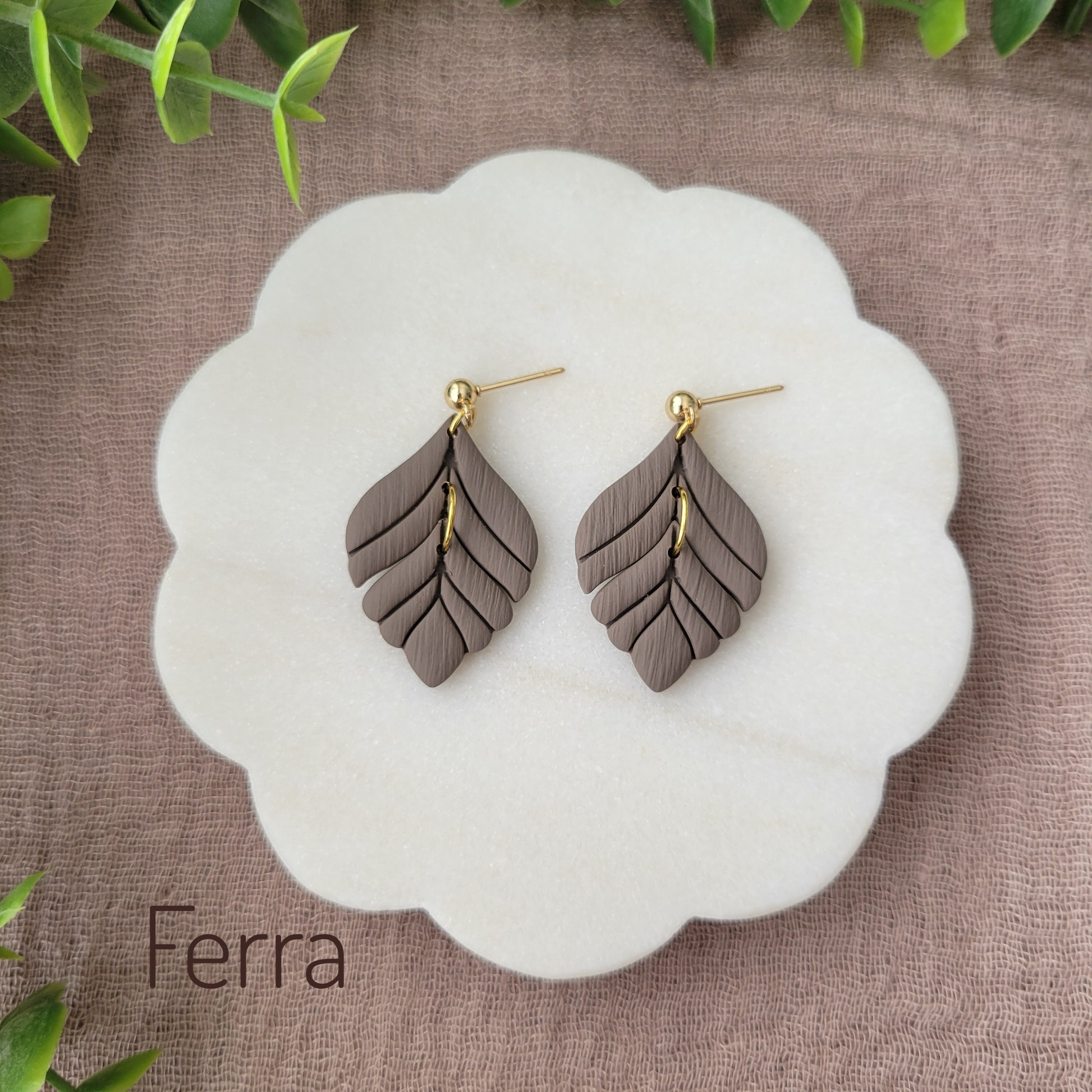 Top view of a pair of greyish brown elegant leaf shaped earrings displayed on a white round scalloped tray.