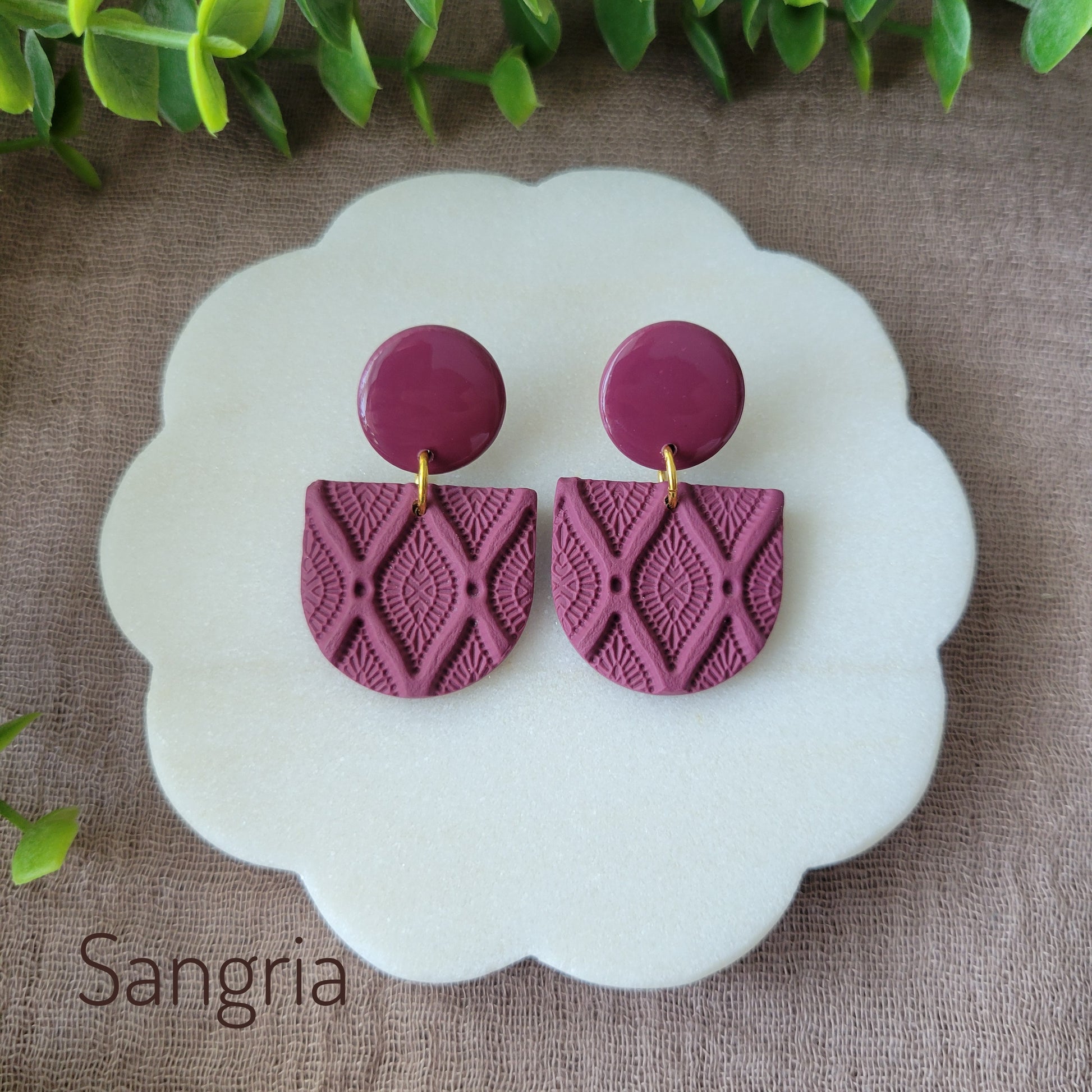 Top view of a pair of reddish purple earrings with deco diamond texture displayed on a white round scalloped tray.