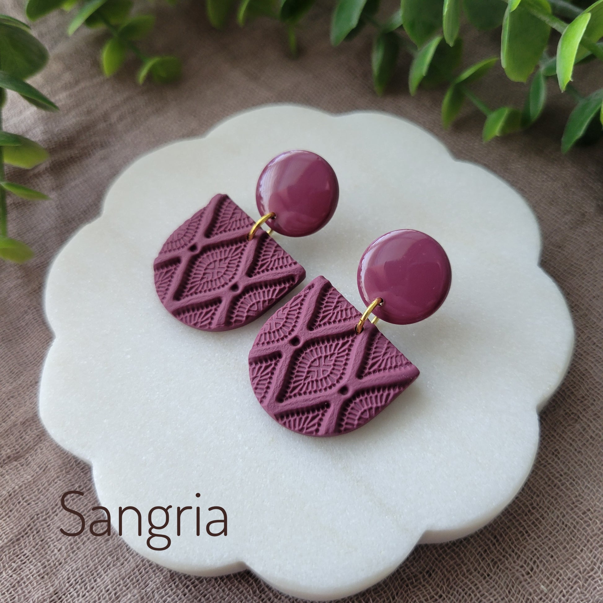 Side view of a pair of reddish purple earrings with deco diamond texture displayed on a white round scalloped tray.