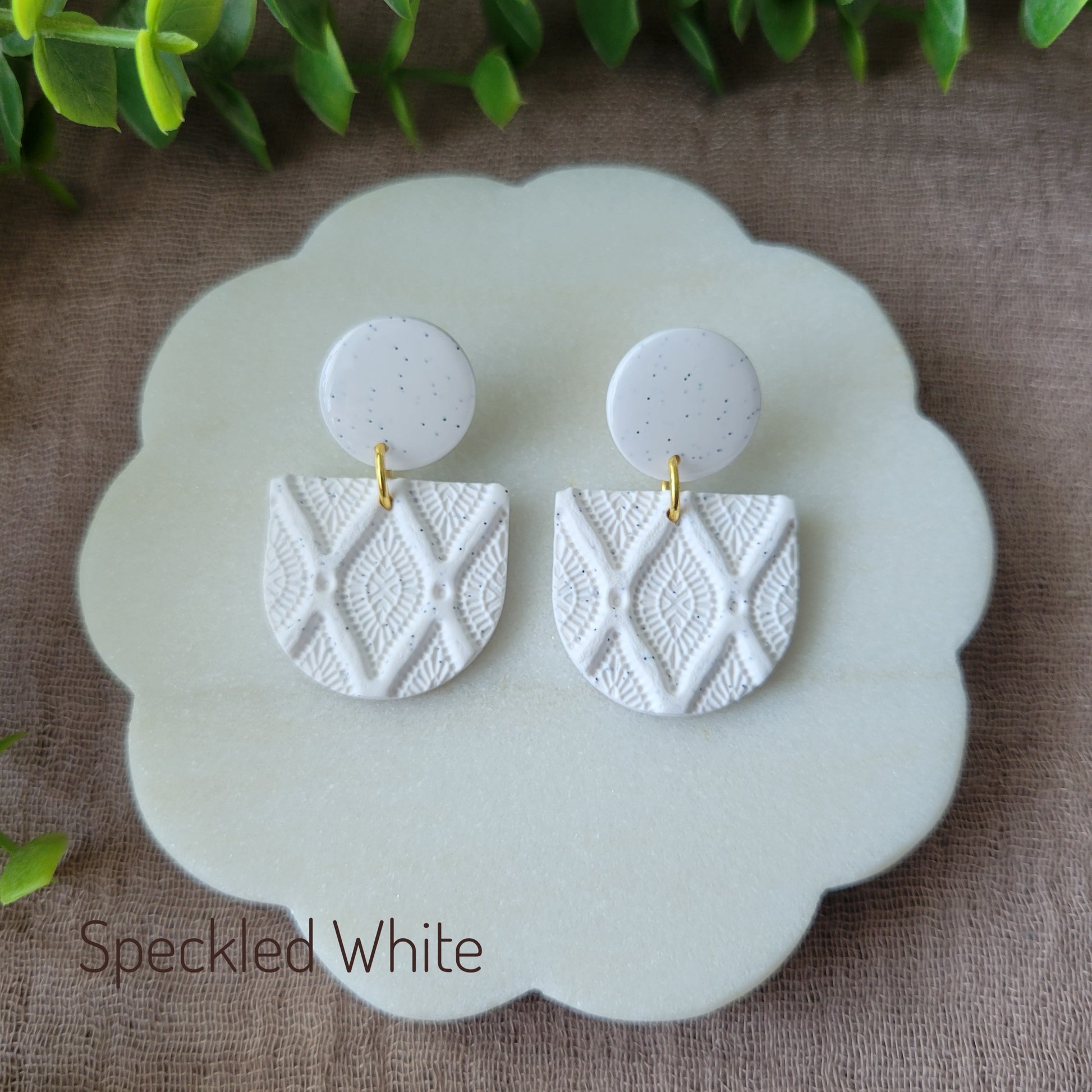 Top view of a pair of speckled white earrings with deco diamond texture displayed on a white round scalloped tray.