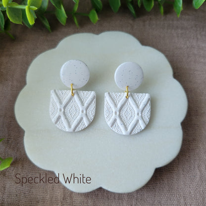 Top view of a pair of speckled white earrings with deco diamond texture displayed on a white round scalloped tray.