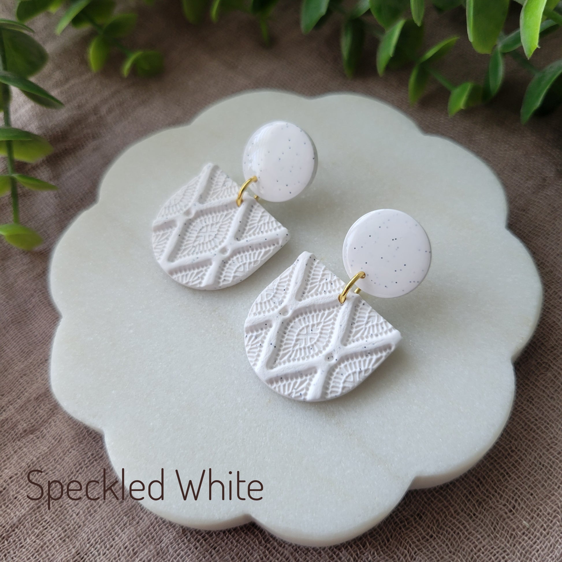 Side view of a pair of speckled white earrings with deco diamond texture displayed on a white round scalloped tray.
