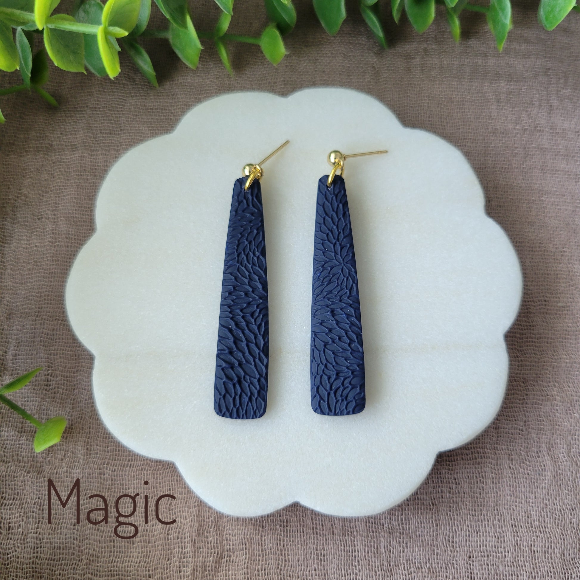 Top view of a pair of dark blue pillar earrings with interwoven leaves texture displayed on a white round scalloped tray.
