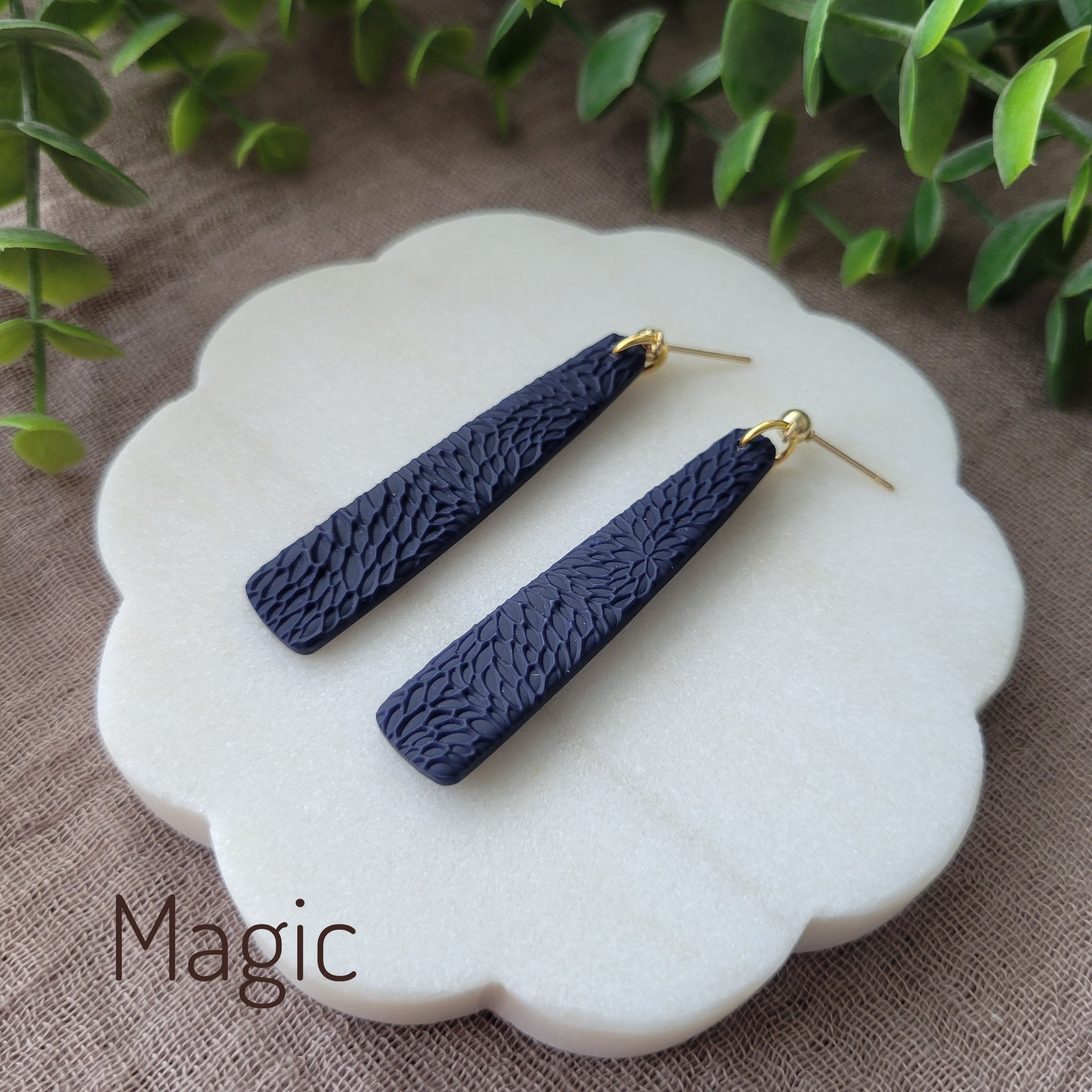 Side view of a pair of dark blue pillar earrings with interwoven leaves texture displayed on a white round scalloped tray.