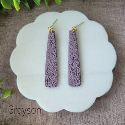 Top view of a pair of light purple pillar earrings with interwoven leaves texture displayed on a round white scalloped tray.