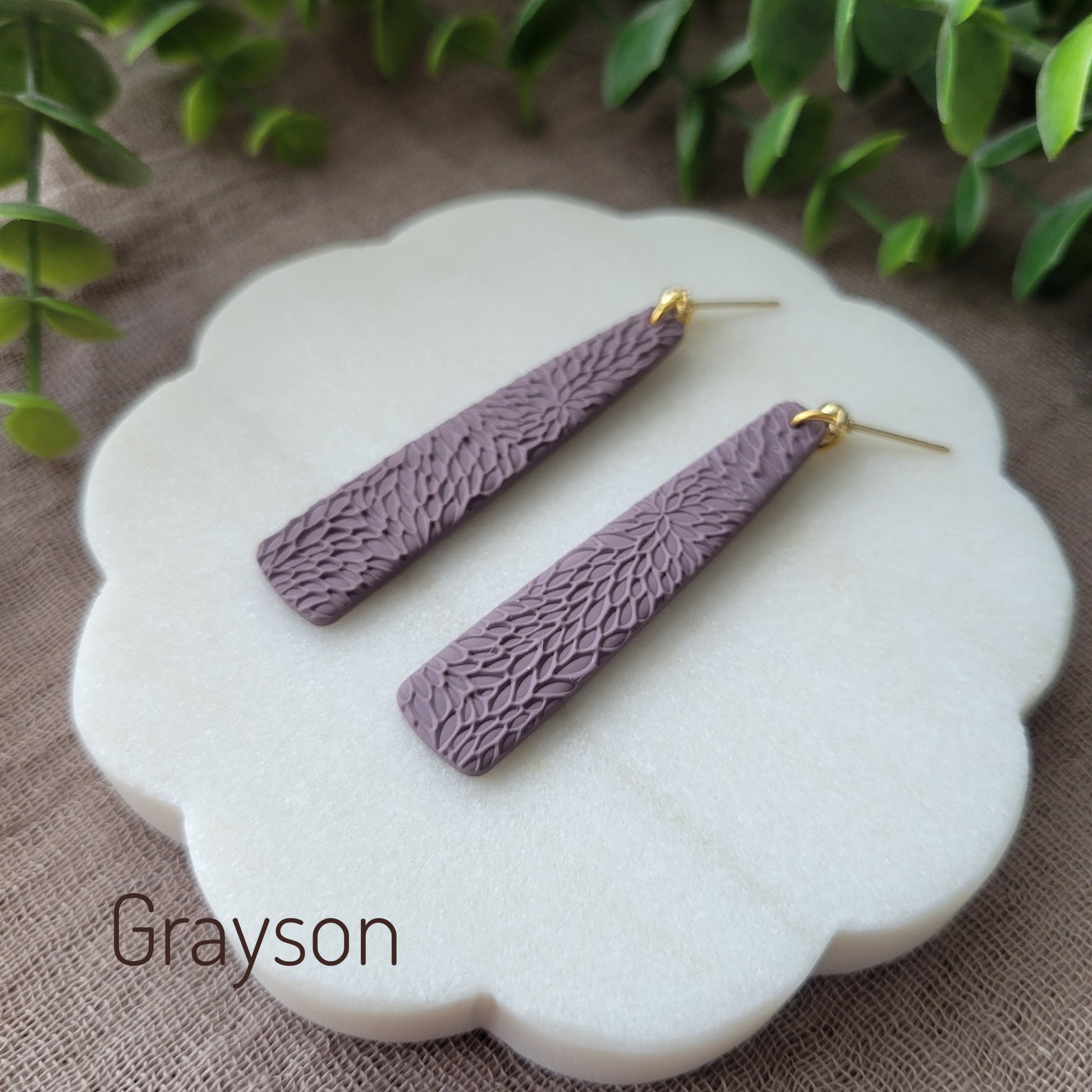 Side view of a pair of light purple pillar earrings with interwoven leaves texture displayed on a white round scalloped tray.
