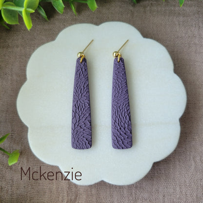 Top view of a pair of dark purple pillar earrings with interwoven leaves texture displayed on a white round scalloped tray.