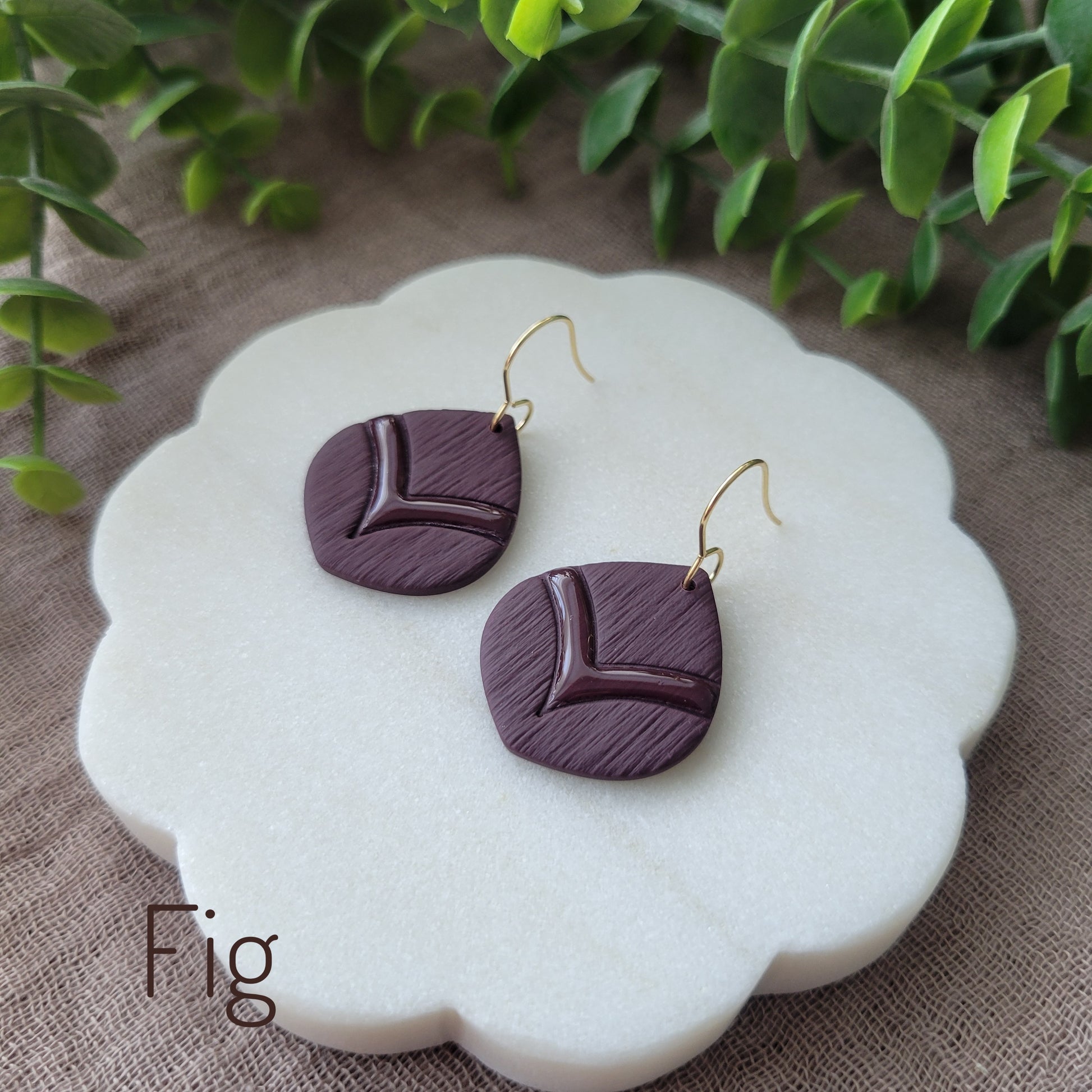 Side view of a pair of dark reddish purple embossed round teardrop earrings displayed on a white round scalloped tray.
