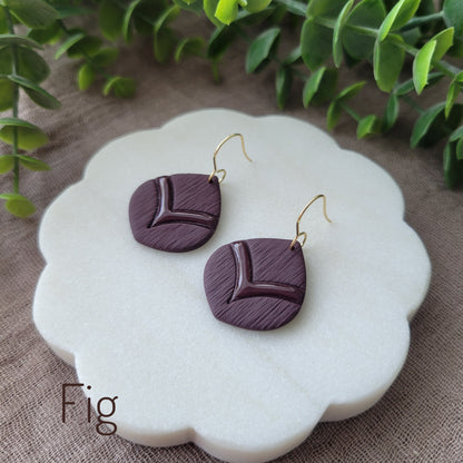 Side view of a pair of dark reddish purple embossed round teardrop earrings displayed on a white round scalloped tray.